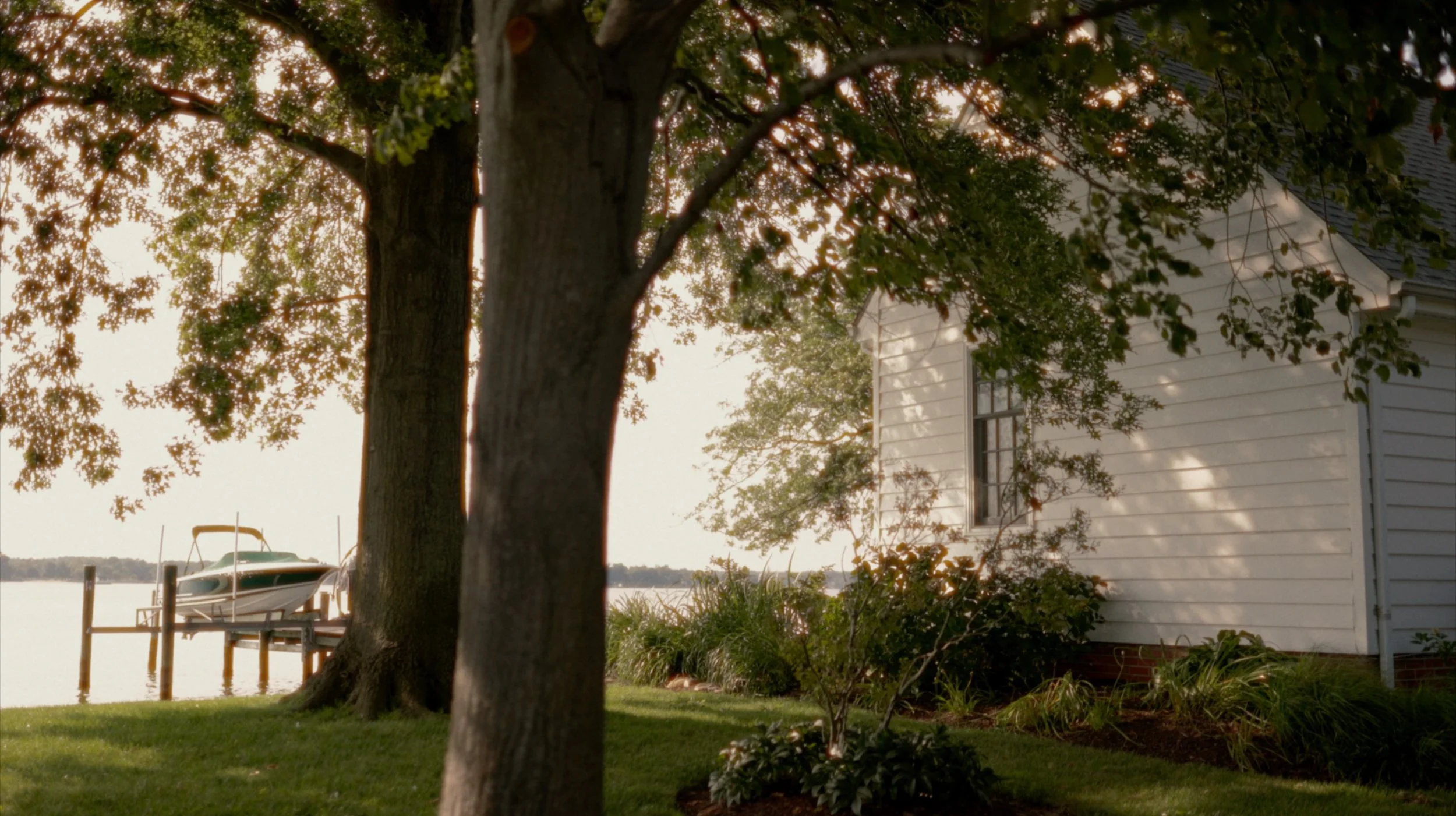 A house by the lake with a dock and boat, trees and greenery in the foreground, and the water in the background.