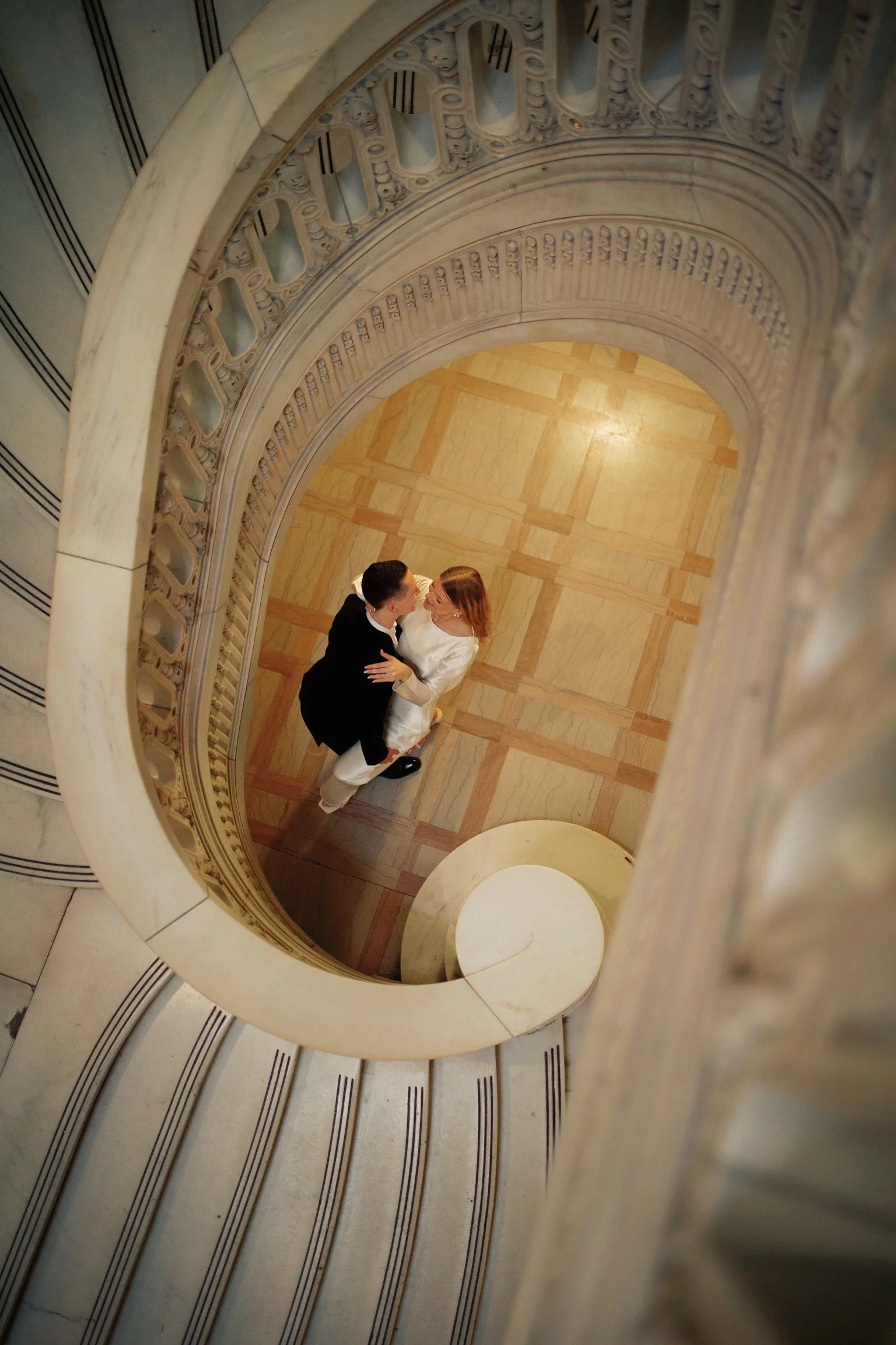 A man in a tuxedo and a woman in a white dress sharing a tender moment on a spiral staircase, viewed from above.