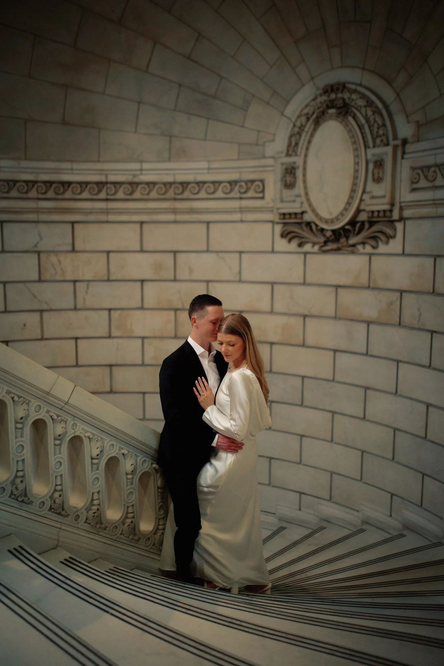 A man and woman share an intimate moment on a grand staircase inside a historic building, with detailed stone walls and decorative architectural elements.