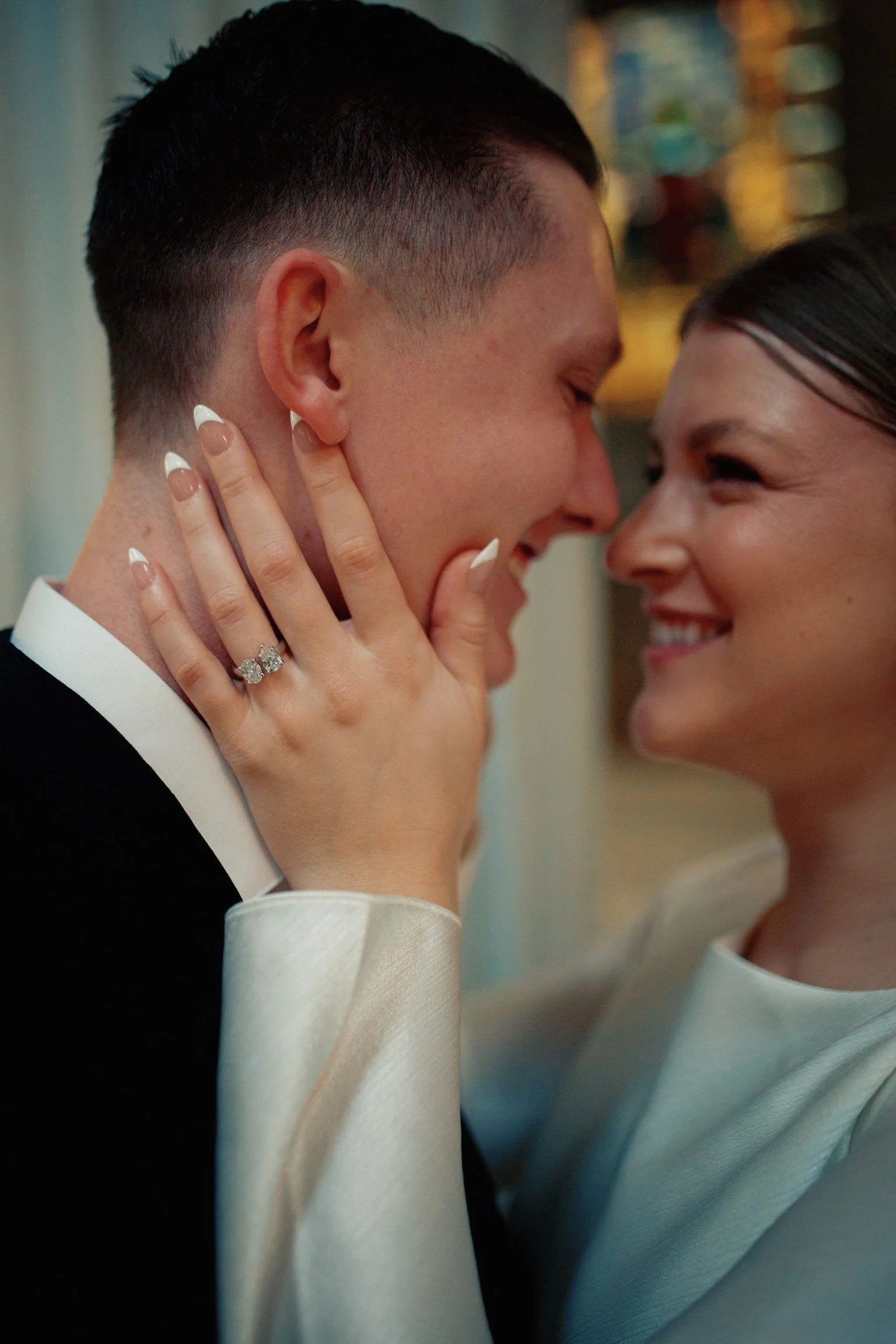 A couple on their wedding day, smiling and holding each other close with their foreheads touching. The woman is wearing a white dress and a ring, and the man is dressed in a black tuxedo.