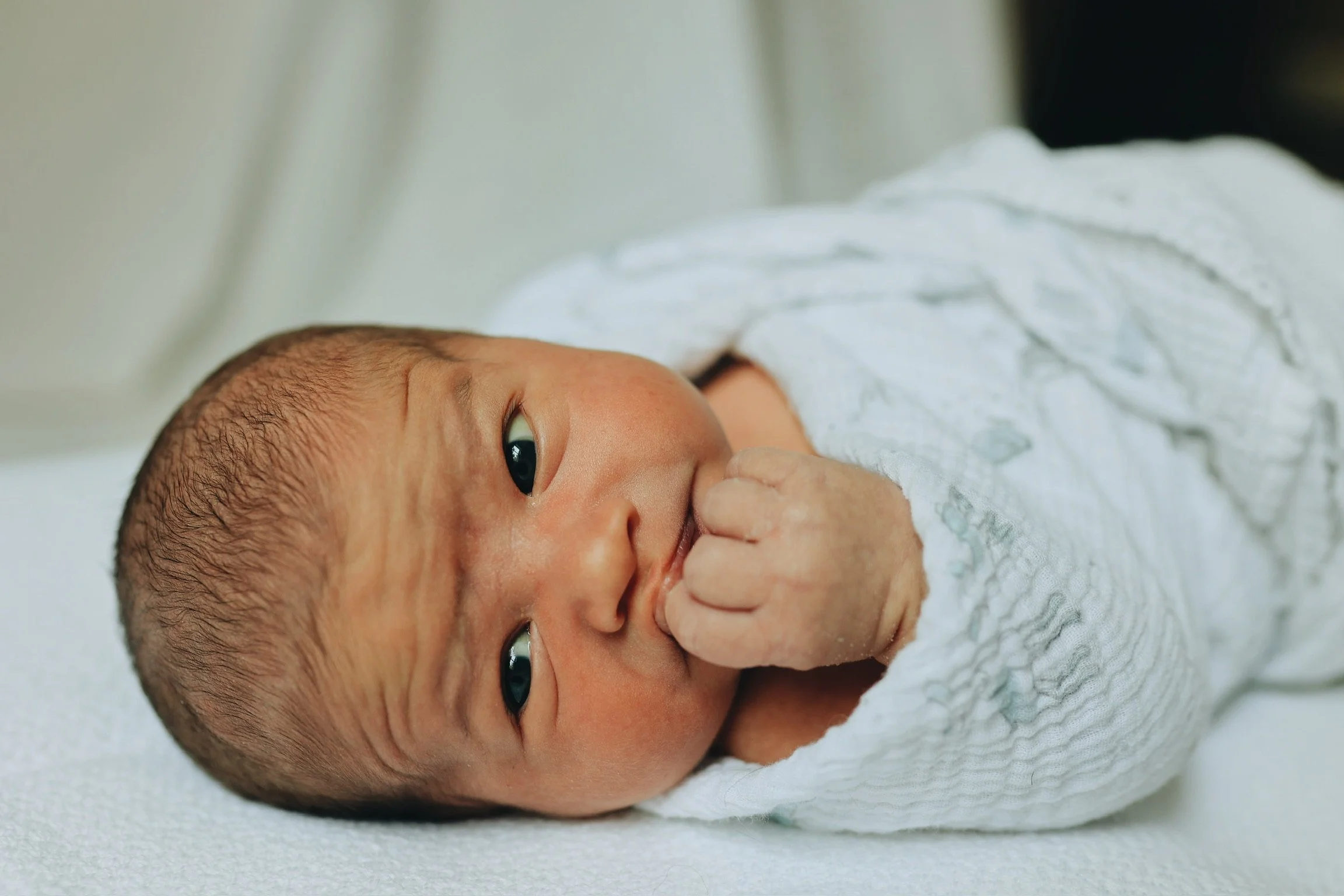 Close-up of an Asian newborn baby lying on a white blanket, wearing a white onesie, with one hand near their mouth, looking directly with wide eyes.