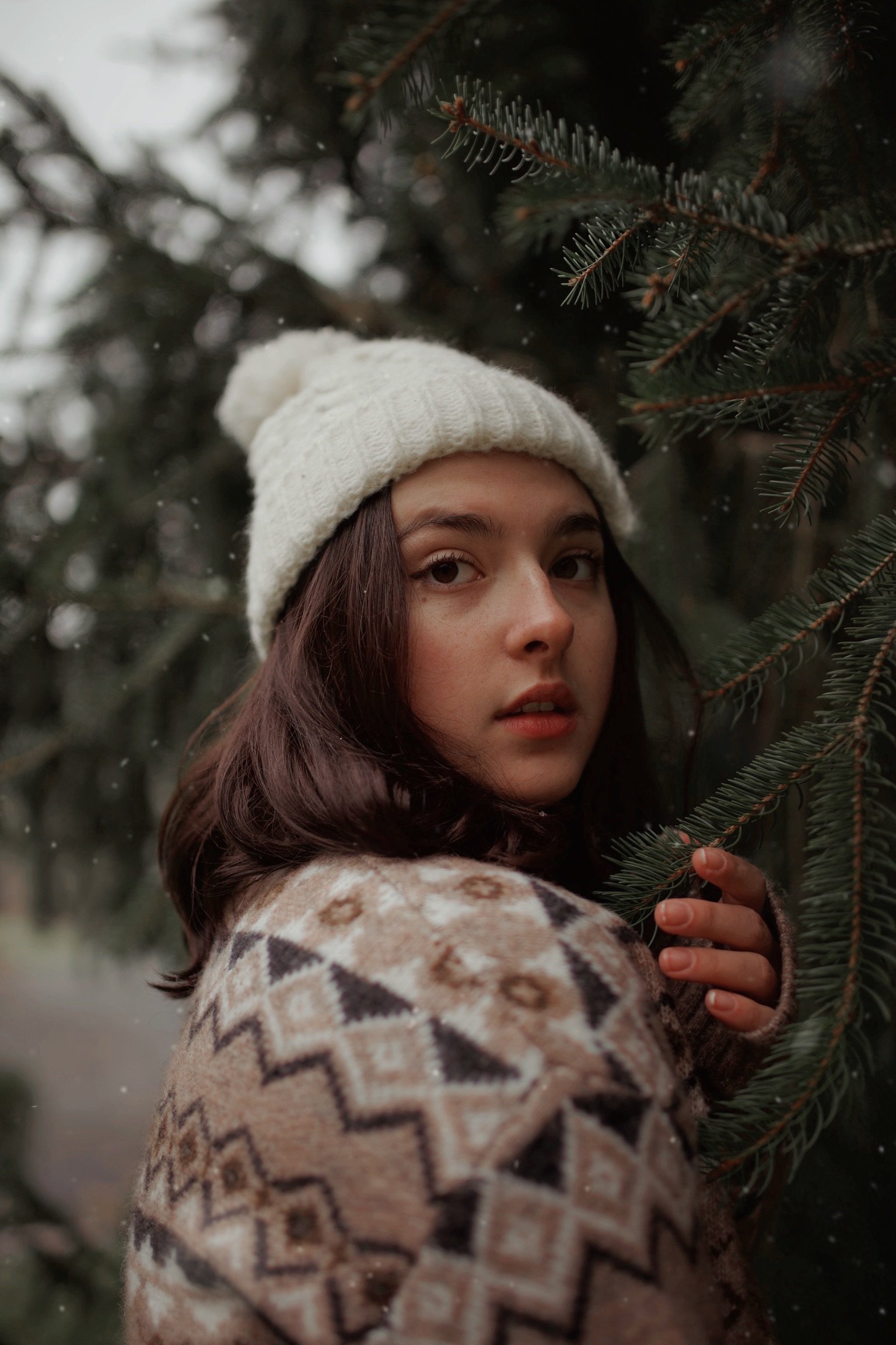 A young woman with dark brown hair wearing a white knit beanie and a patterned sweater, standing outdoors among evergreen trees, holding a branch of the tree.