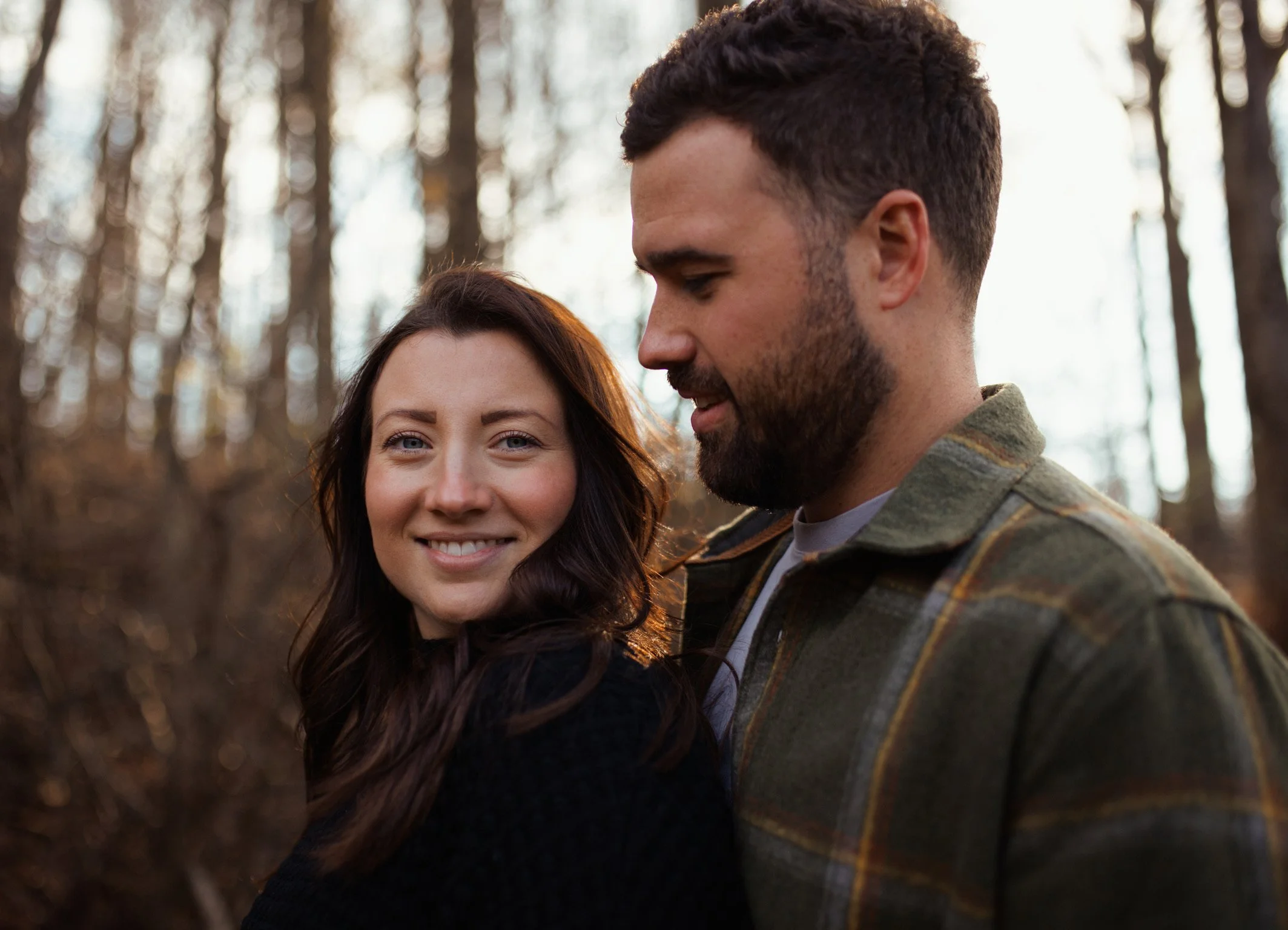A smiling woman with long brown hair and a man with a beard looking down at her, standing in a forest during sunset.
