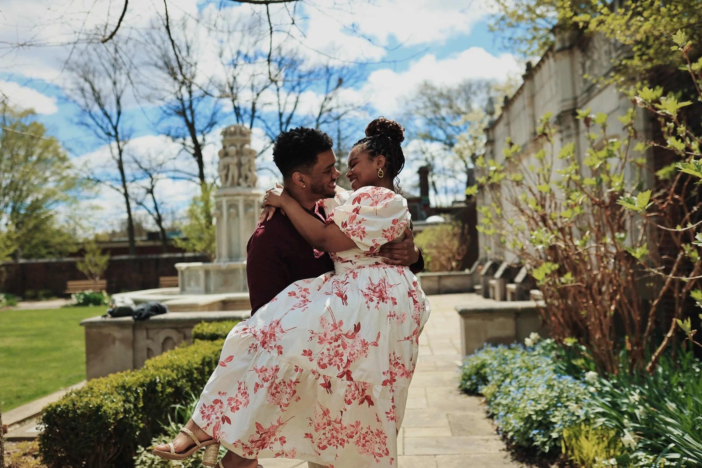 A happy couple embracing outdoors in a garden with a fountain, trees, and a building in the background on a bright, partly cloudy day.