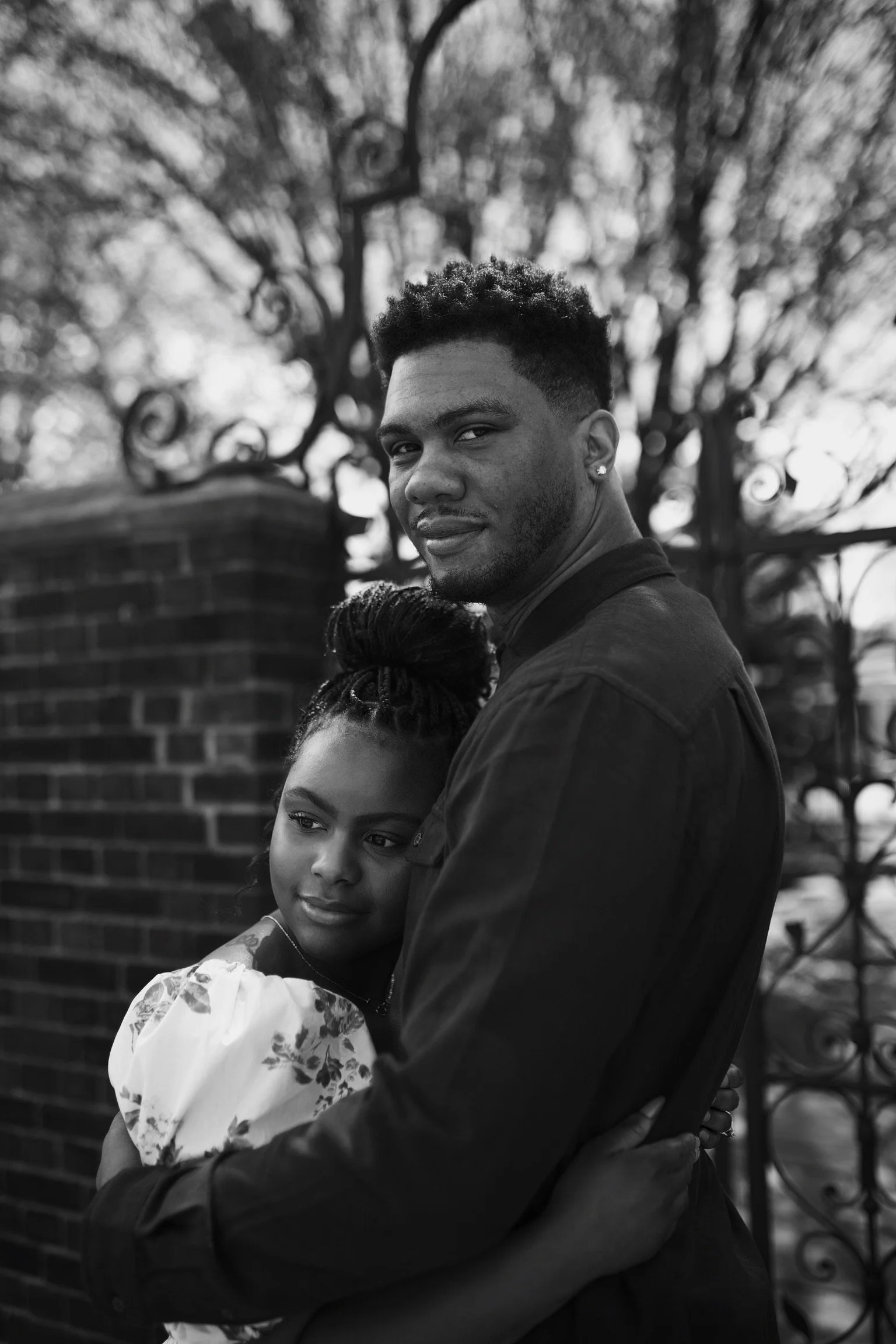 A black and white photo of a couple standing close, with the woman resting her head on the man's chest, both looking relaxed and happy. The man has short hair and an earring, while the woman has braided hair styled up.