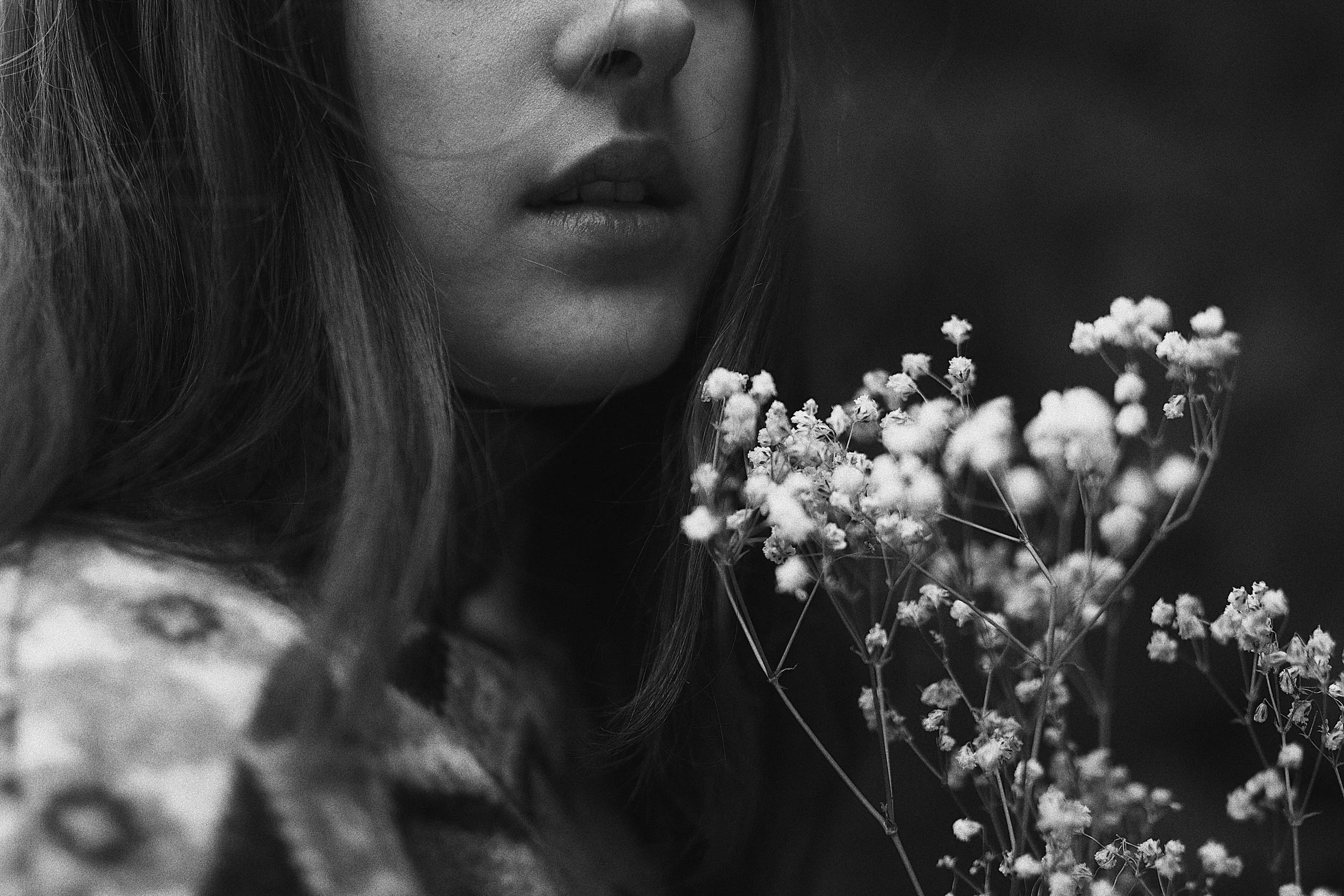 Close-up black and white photo of a woman's face, showing lips, chin, and part of her nose, with long hair. She is holding a cluster of small flowers close to her face.