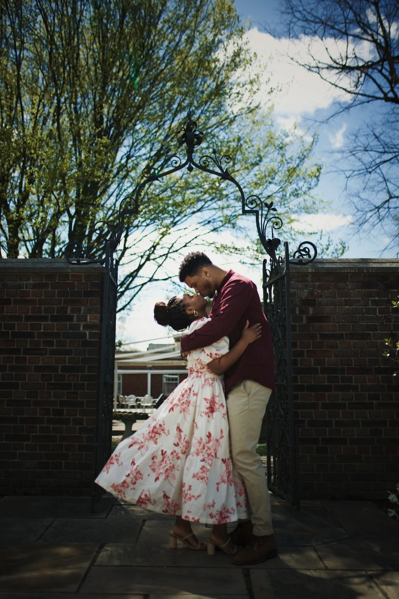 A couple sharing a kiss under a wrought iron gate, with green trees and a blue sky in the background.