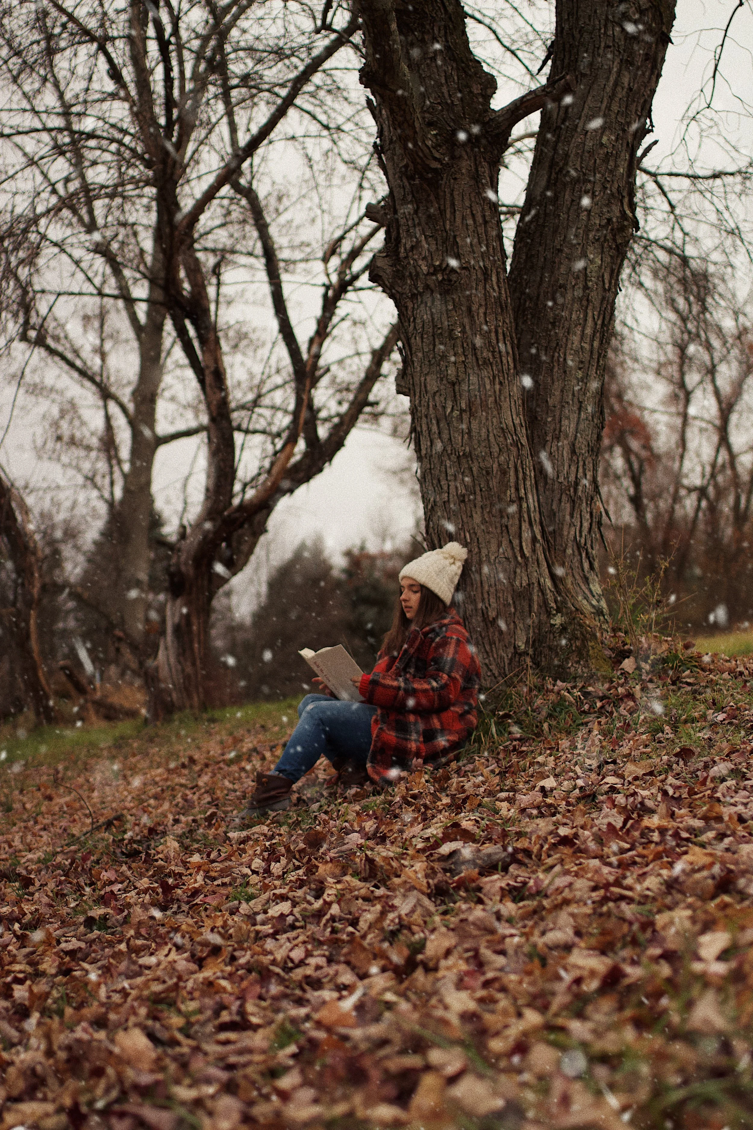 A young woman sitting on the ground leaning against a large tree, reading a book in a leaf-covered park during fall or winter, wearing a knit hat and a plaid coat.