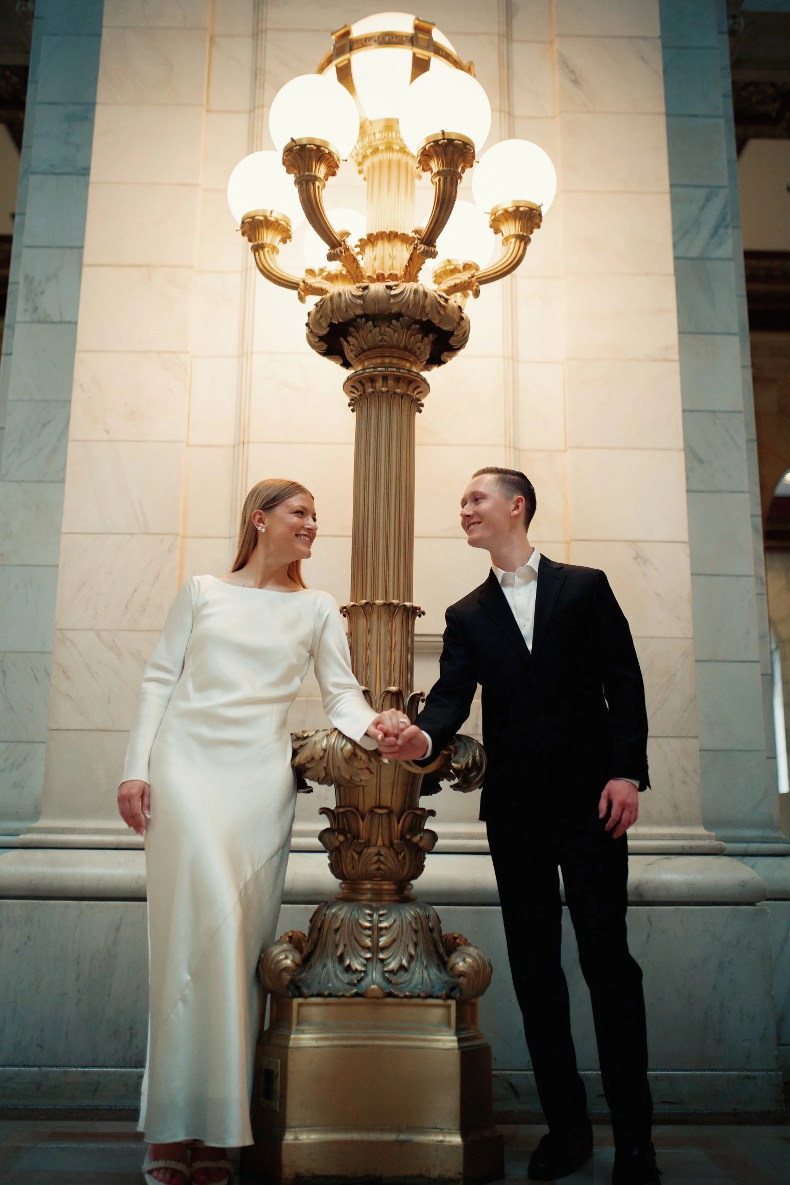 A couple dressed in formal attire, standing hand in hand, smiling and looking at each other near a large ornate streetlamp inside a grand building with marble walls.
