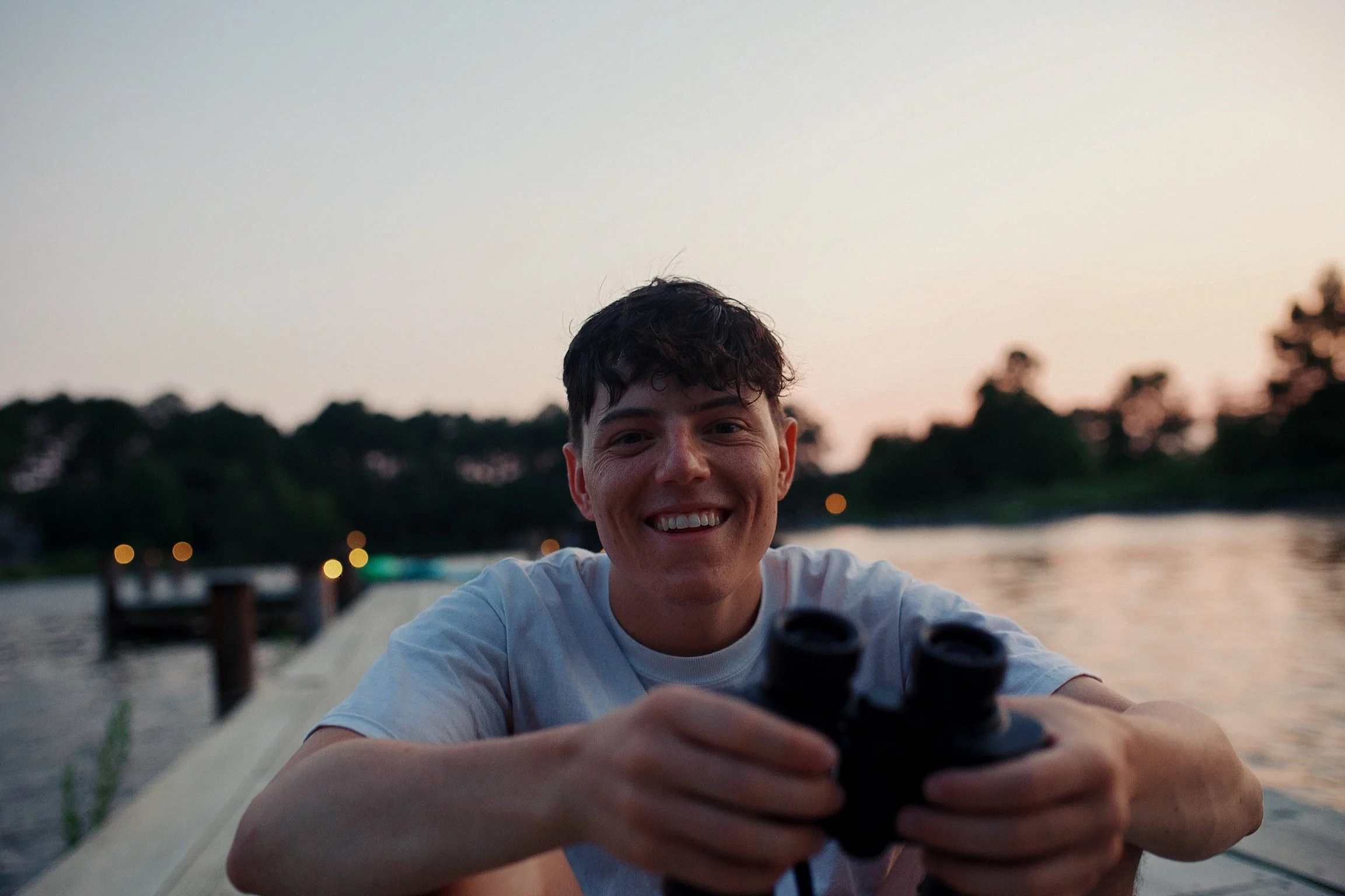 Young man smiling while holding binoculars on a dock by the water at sunset.