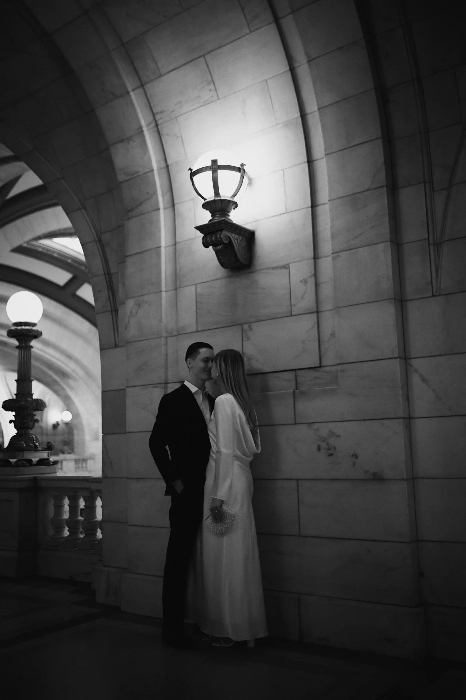 A black-and-white photo of a couple in formal attire standing closely and smiling, inside a historic building with stone walls, arches, and wall-mounted lamps. The woman is wearing a long dress and holding a small purse, while the man is wearing a da