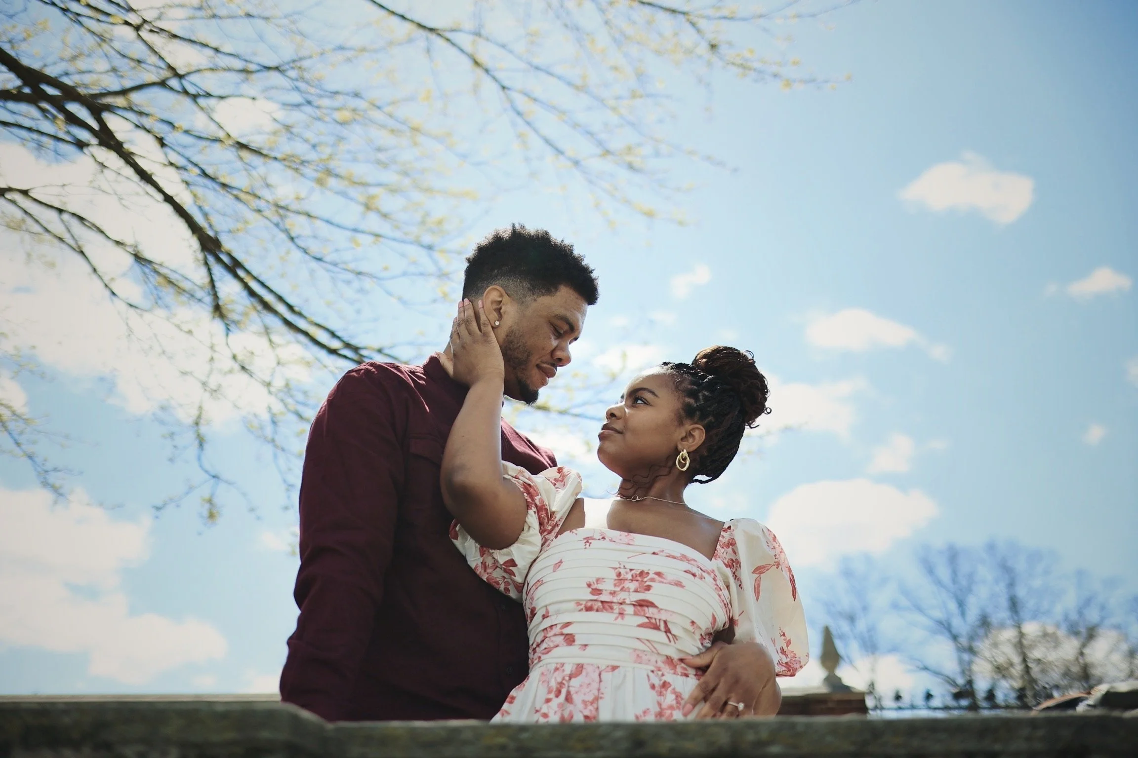 A couple standing outdoors under a blue sky with clouds, looking into each other's eyes, with the woman's hand on the man's neck.