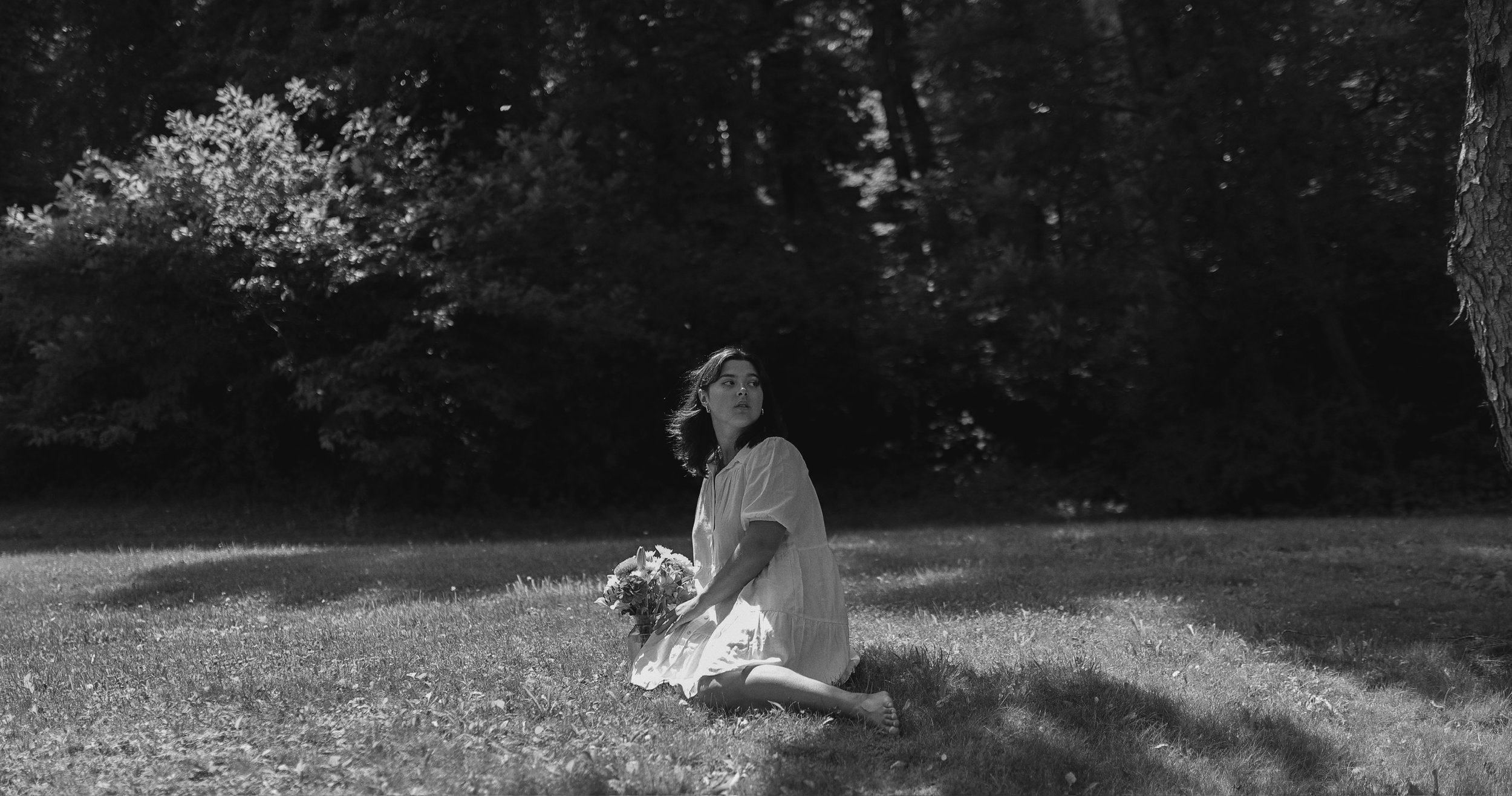 A woman sitting on the grass in a park, holding a bouquet of flowers, with trees and shadows in the background, in black and white.