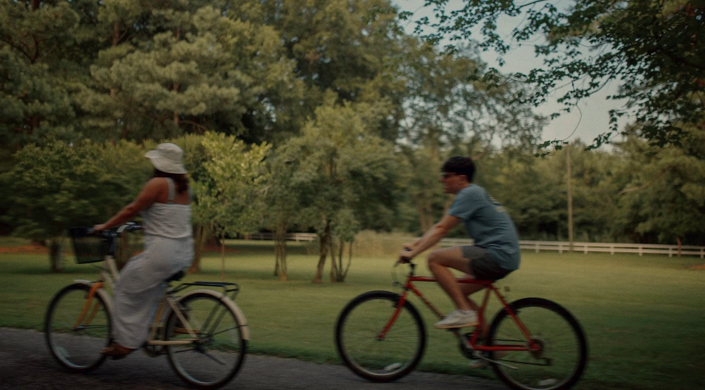 Two people riding bicycles on a path through a park with green trees and grass in the background.