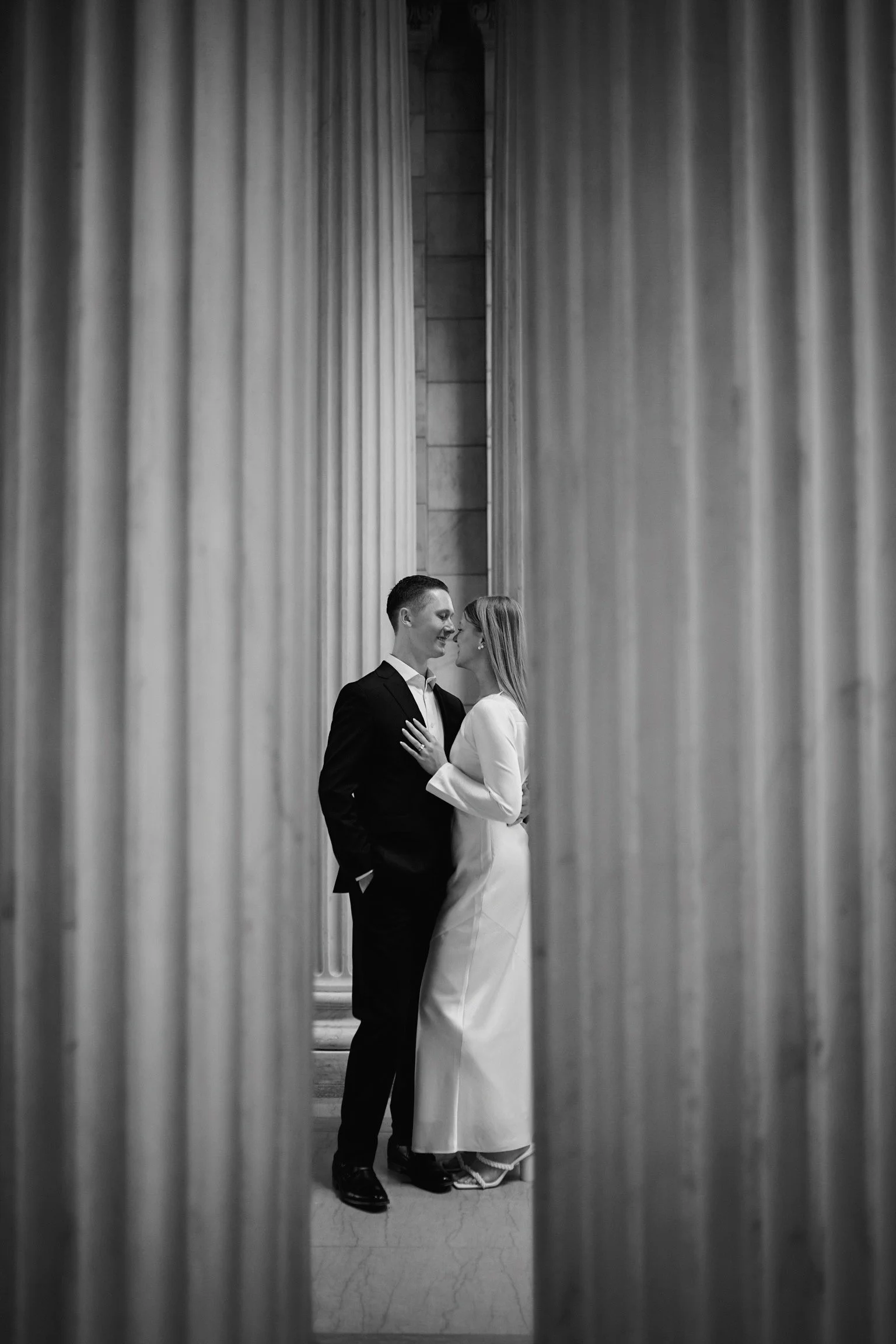 A black and white photo of a couple, a man in a black suit and a woman in a white dress, standing close together between large columns, looking at each other and smiling.