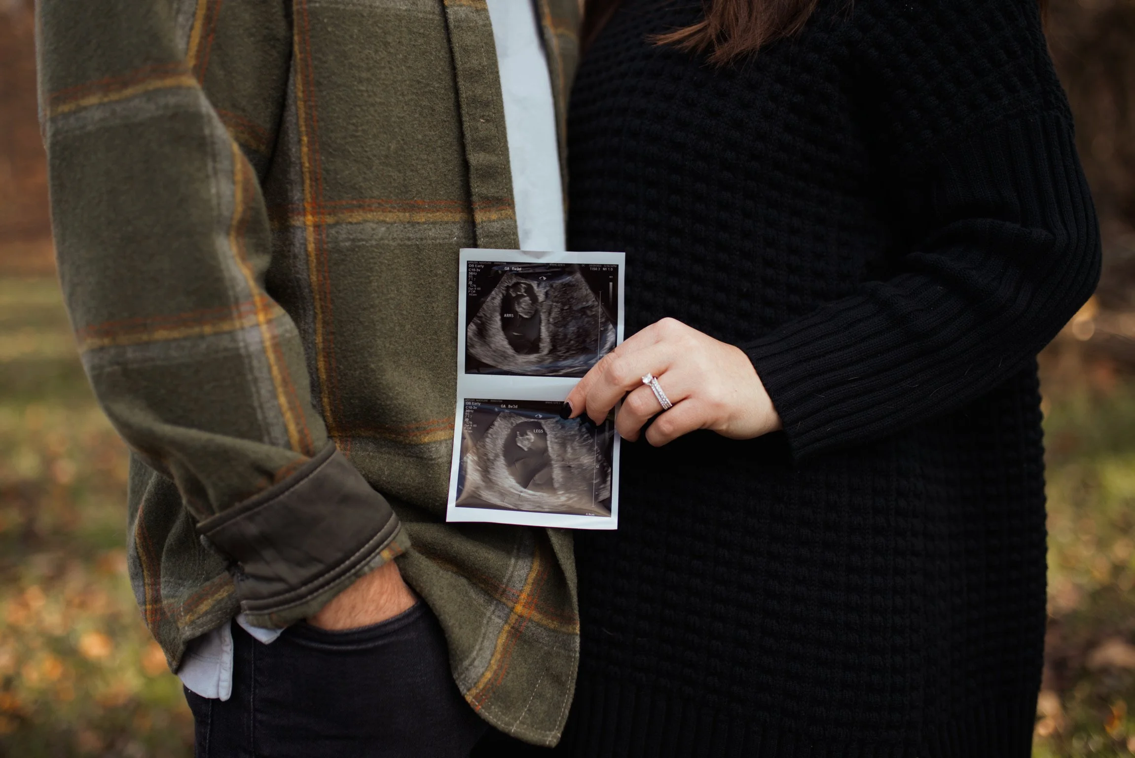 A couple holding ultrasound images of a fetus outdoors, with the woman wearing a ring on her finger.