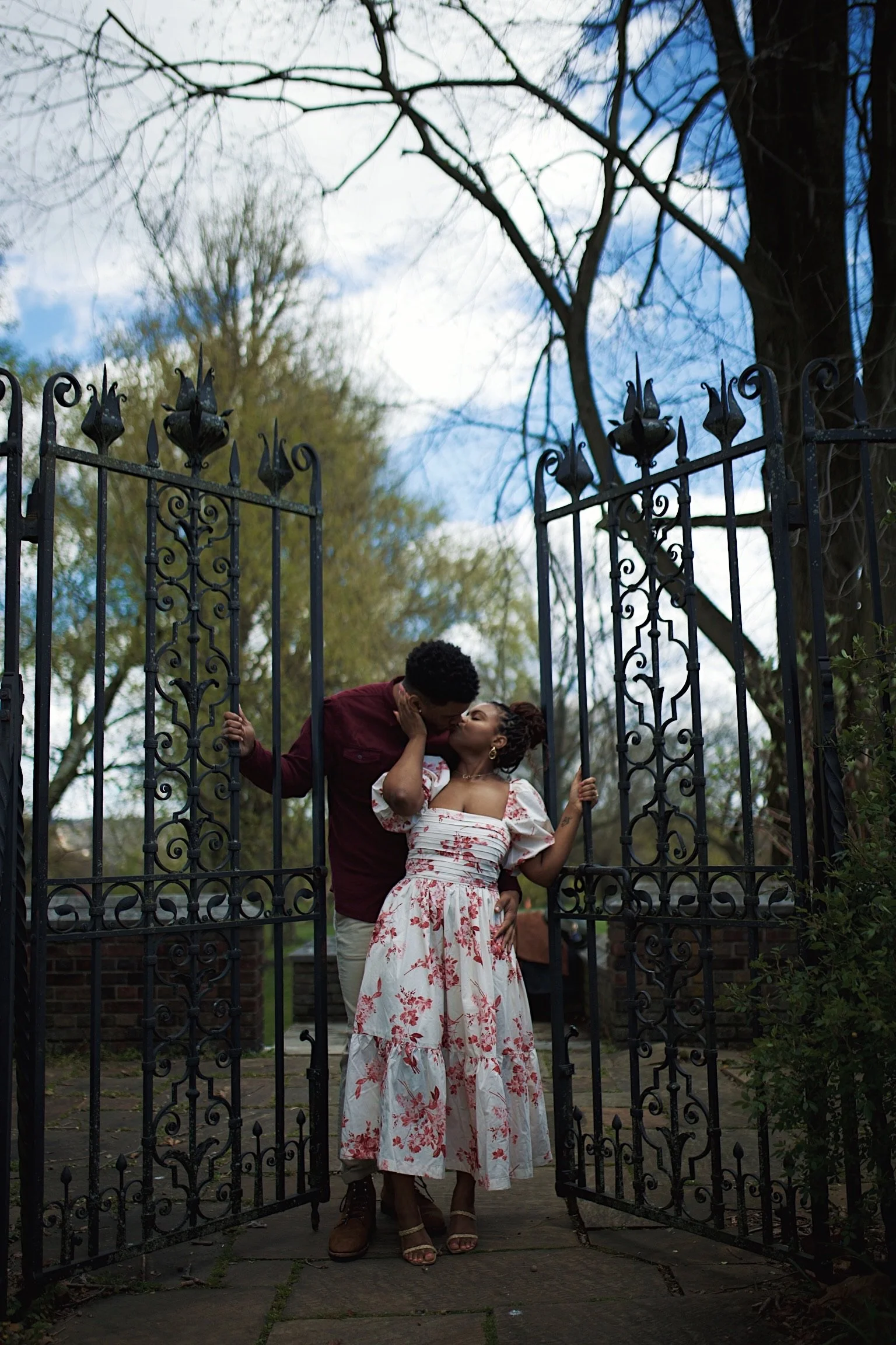 A couple sharing a kiss at an open wrought iron gate, with trees and a partly cloudy sky in the background.