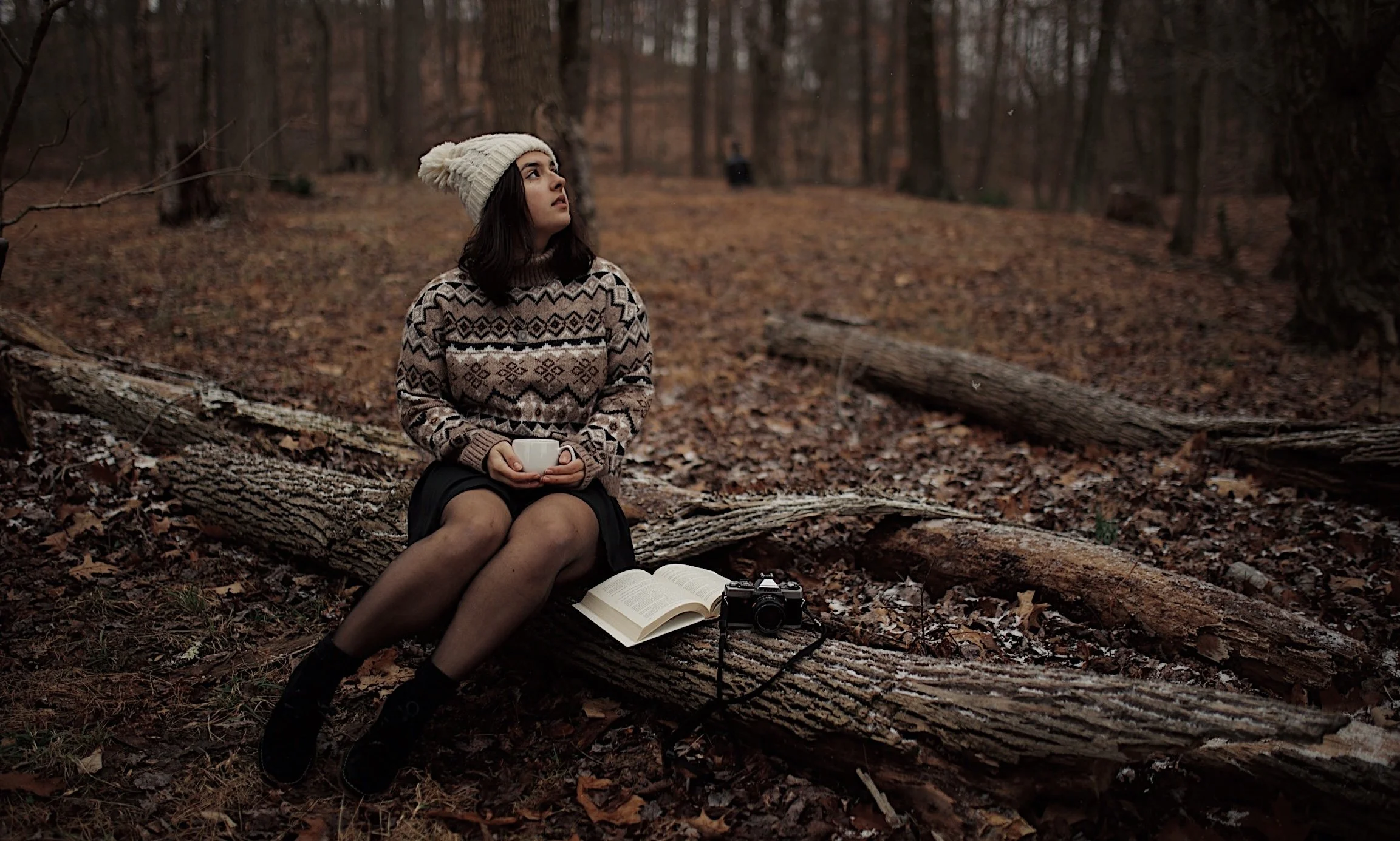 Young woman sitting on a fallen log in a forest during autumn, holding a cup, with an open book and camera beside her.
