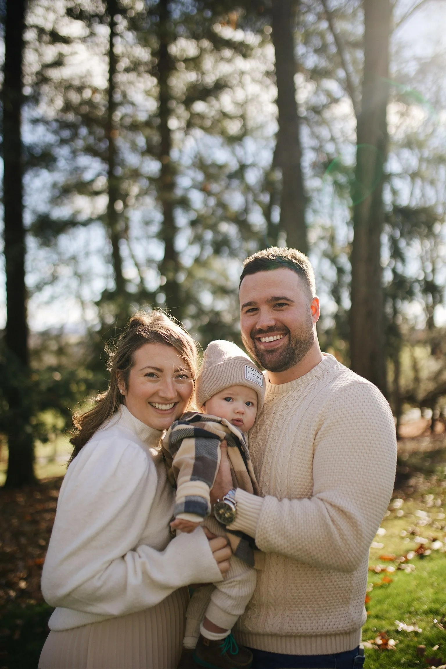 A smiling family of three, a woman, a man, and a baby, enjoying time outdoors in a forest on a sunny day.