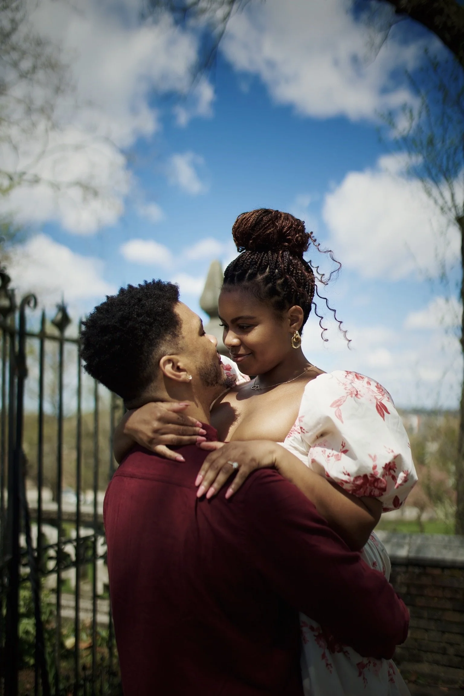 A couple celebrating an engagement, with the man holding the woman in his arms as they look into each other's eyes outdoors under a blue sky with clouds.