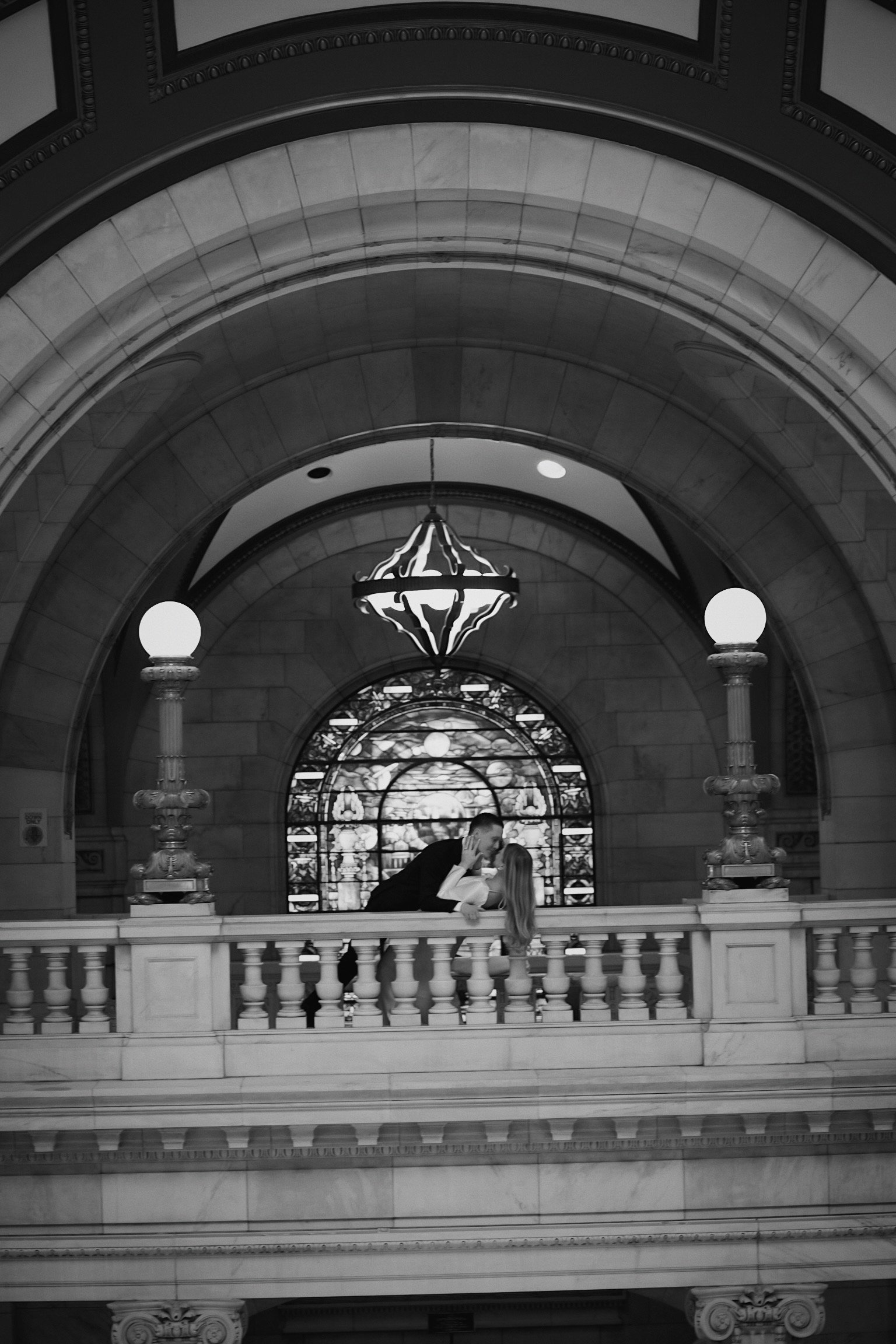 A black and white photo of a man and woman kissing on a balcony inside a grand building with arched ceilings, ornate light fixtures, and stained glass windows.