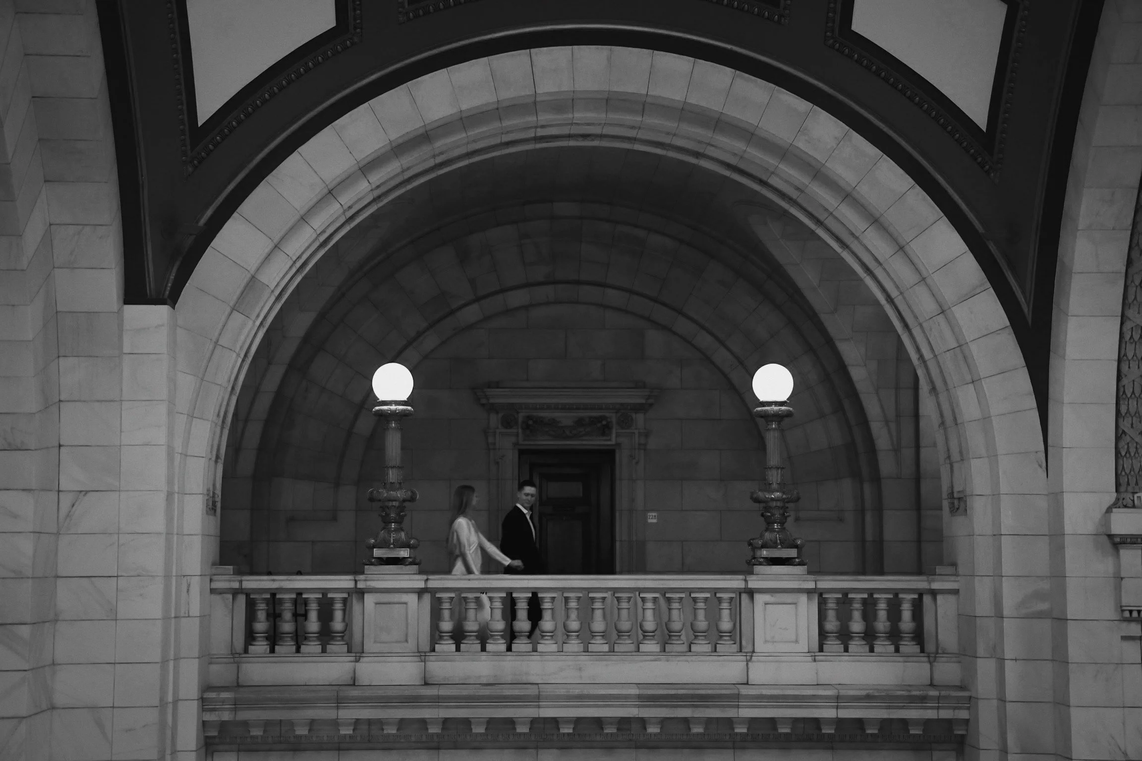 Two people, a woman and a man, holding hands on a marble balcony inside a large historic building with arched windows and ornate lamps.