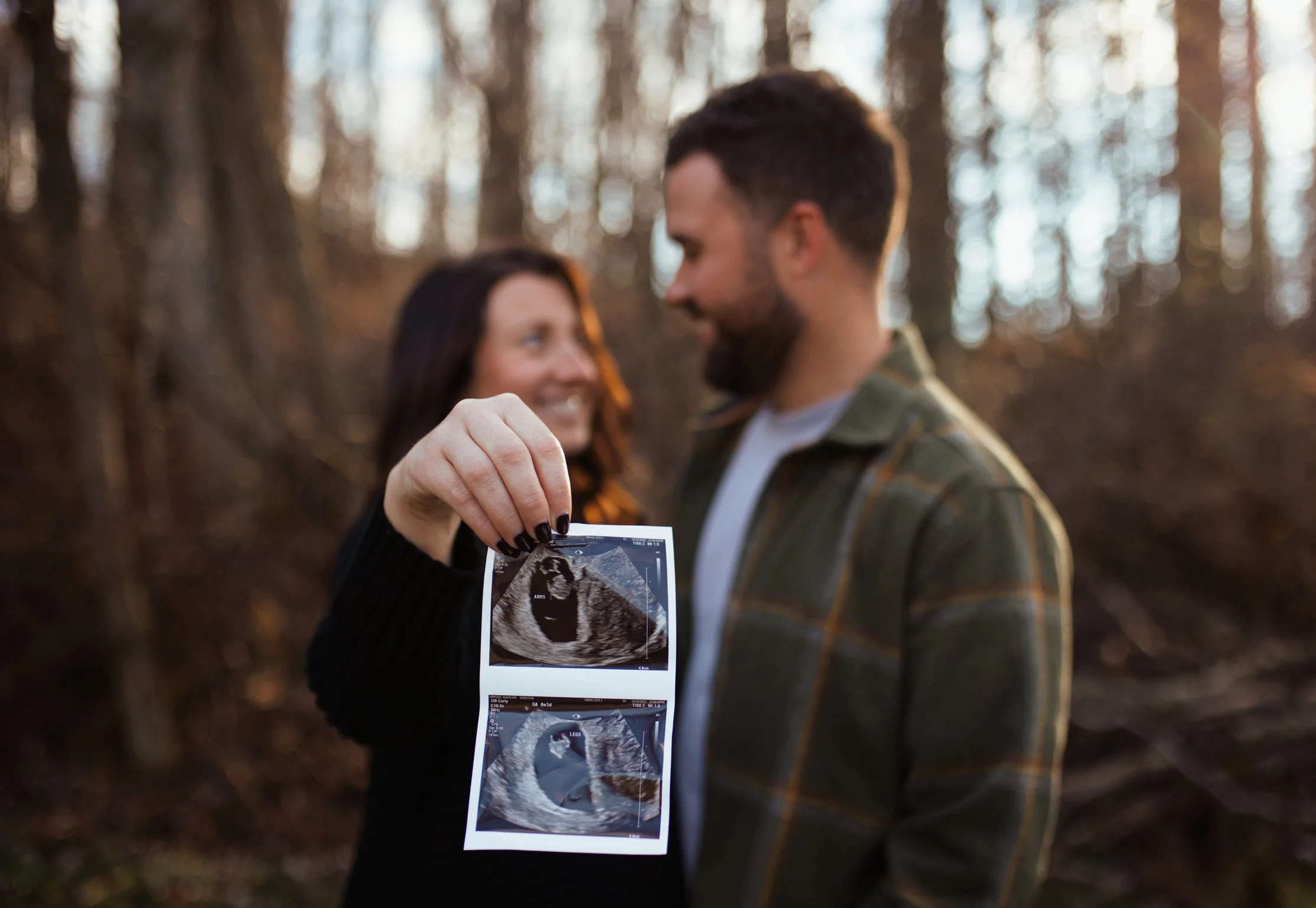 A couple stands in a wooded outdoor area during sunset or late afternoon, with the woman holding ultrasound pictures of a fetus, both smiling and looking at each other.