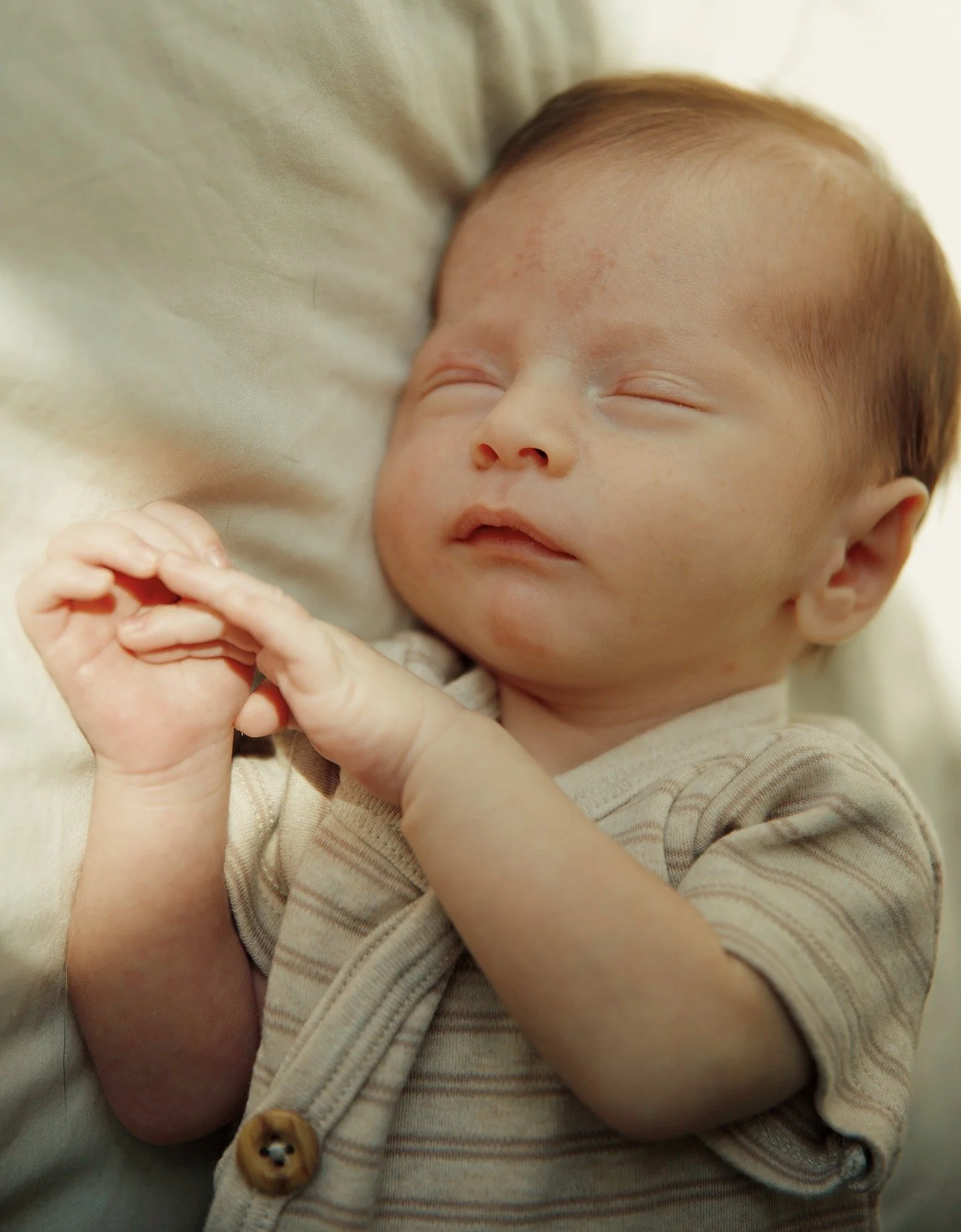 A sleeping baby with light skin, short light brown hair, and closed eyes, lying on a soft surface, wearing a beige and brown striped shirt with a small button.