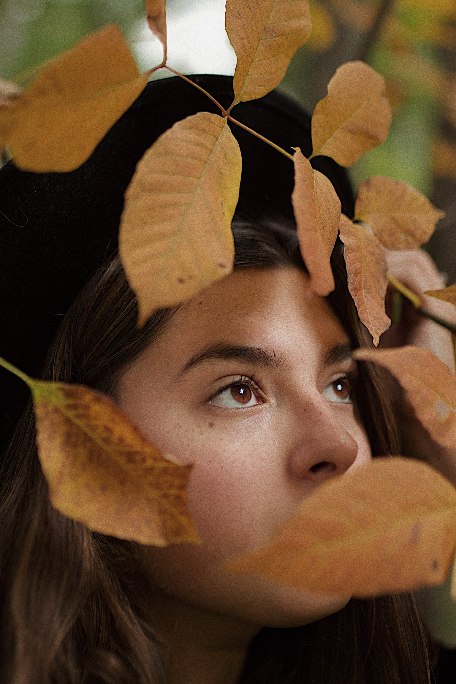 A young woman with brown hair and brown eyes, surrounded by autumn leaves, looking up.