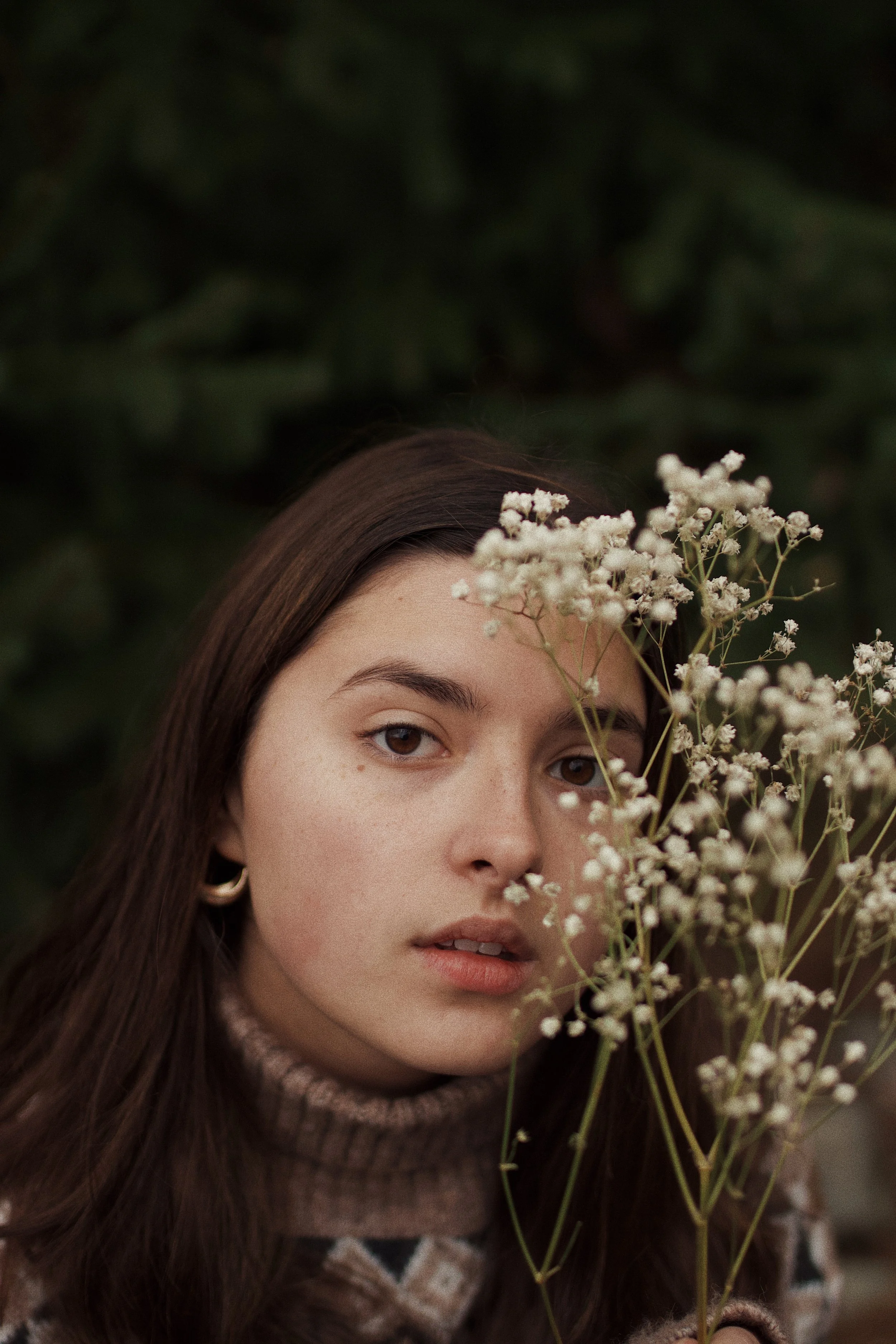 A young woman with long brown hair and hoop earrings gazing at the camera, partially covered by a cluster of small white flowers, with a dark green leafy background.