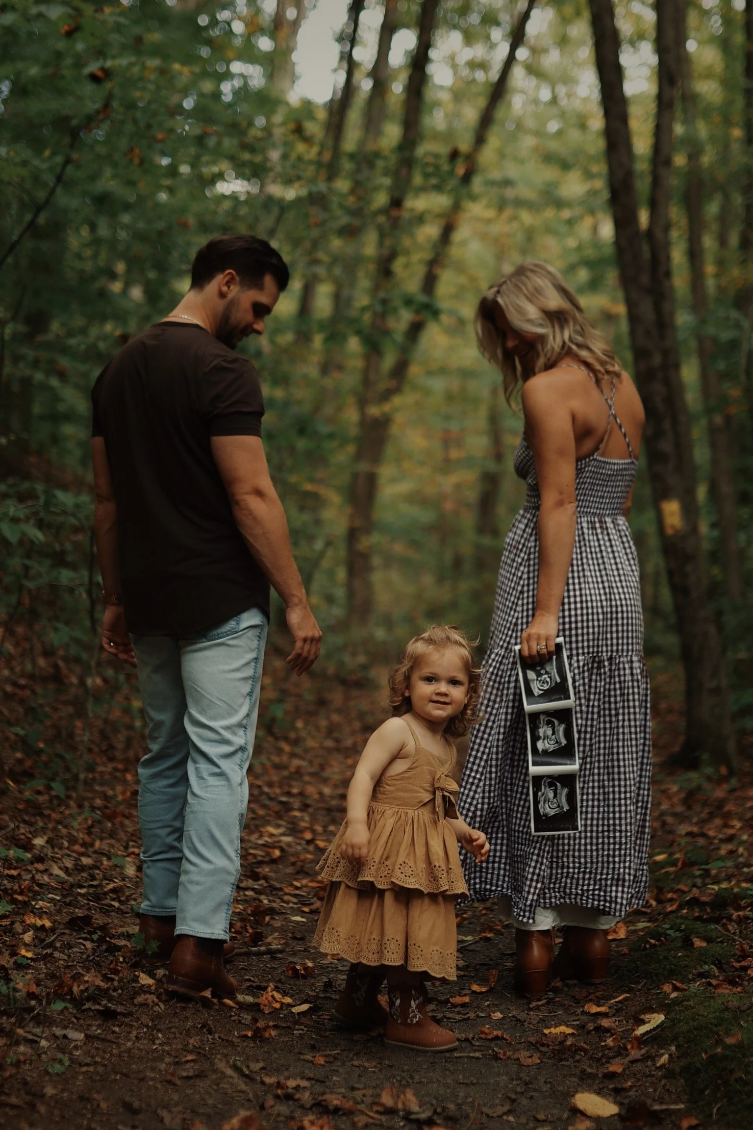 A family of three, including a young girl, standing on a forest trail. The young girl is holding a series of ultrasound images. The adults, a man and a woman, are smiling and looking at the girl. The setting appears to be during autumn.