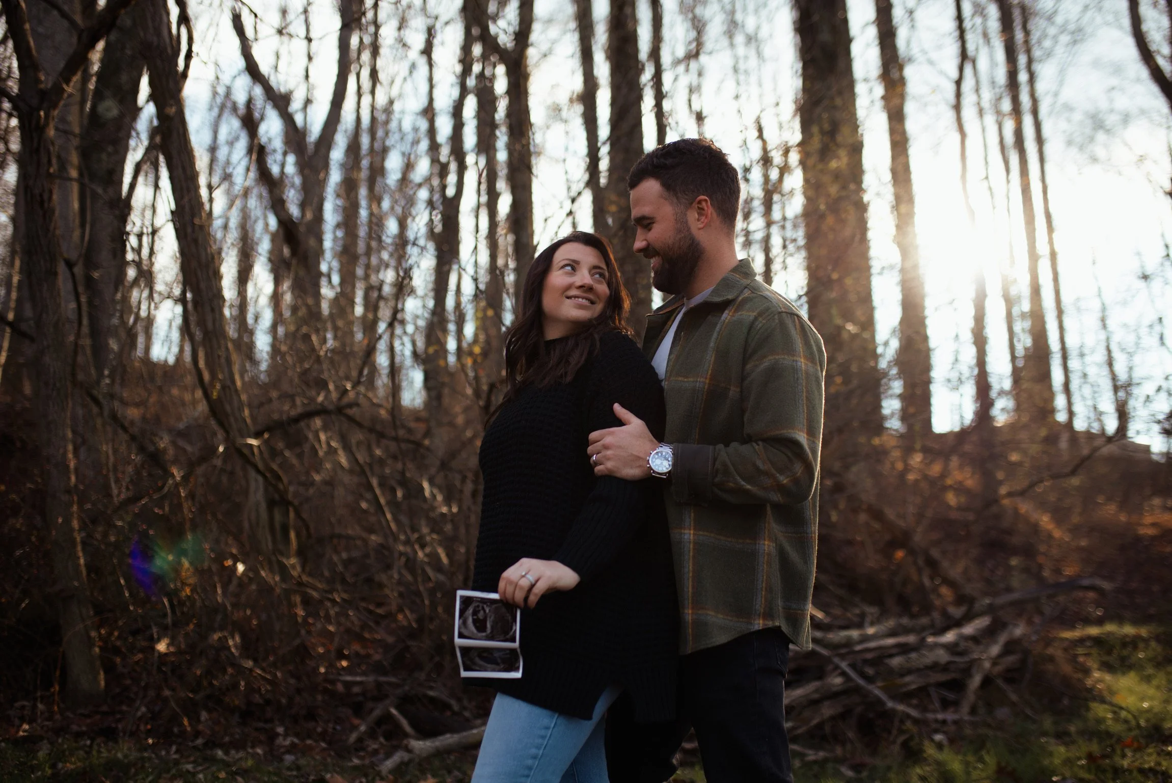 A couple in a forest, with the woman holding an ultrasound image, smiling at each other.
