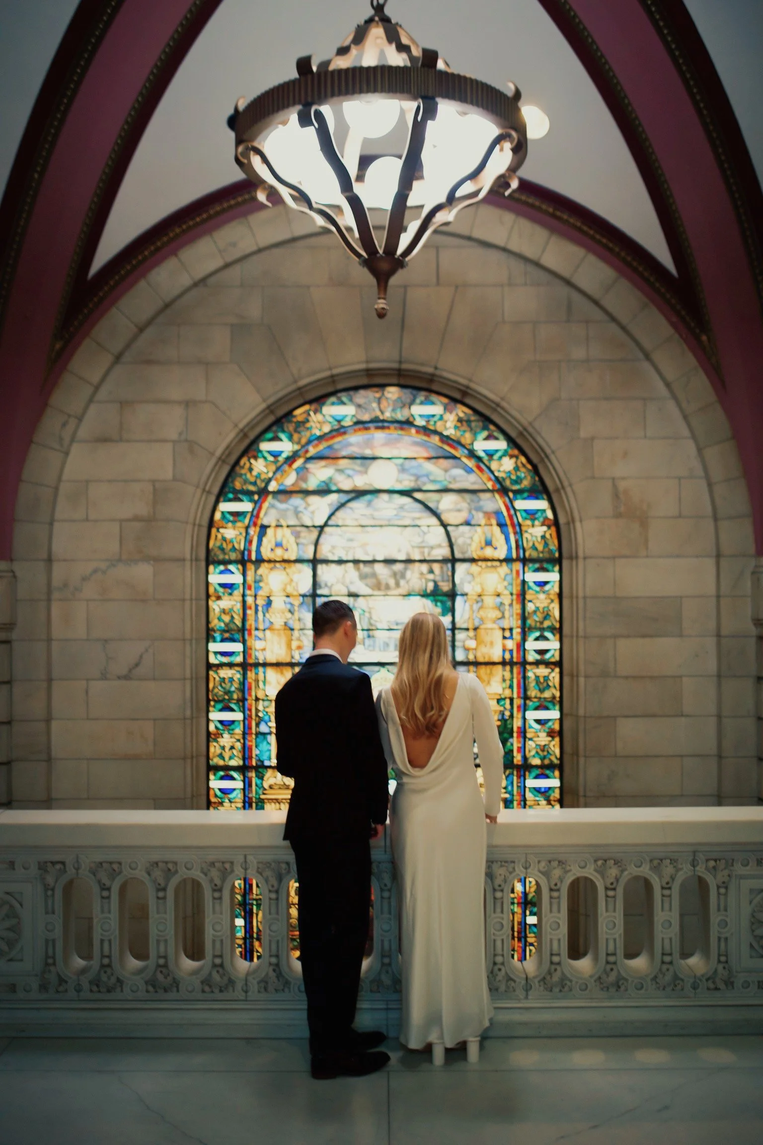 A couple dressed in wedding attire standing together inside a building with a stained glass window behind them.