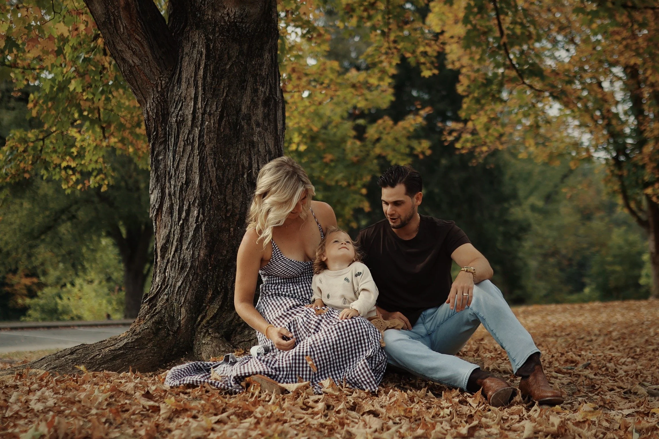 A family of three sitting on autumn leaves under a large tree, with fall foliage in the background, during the daytime.