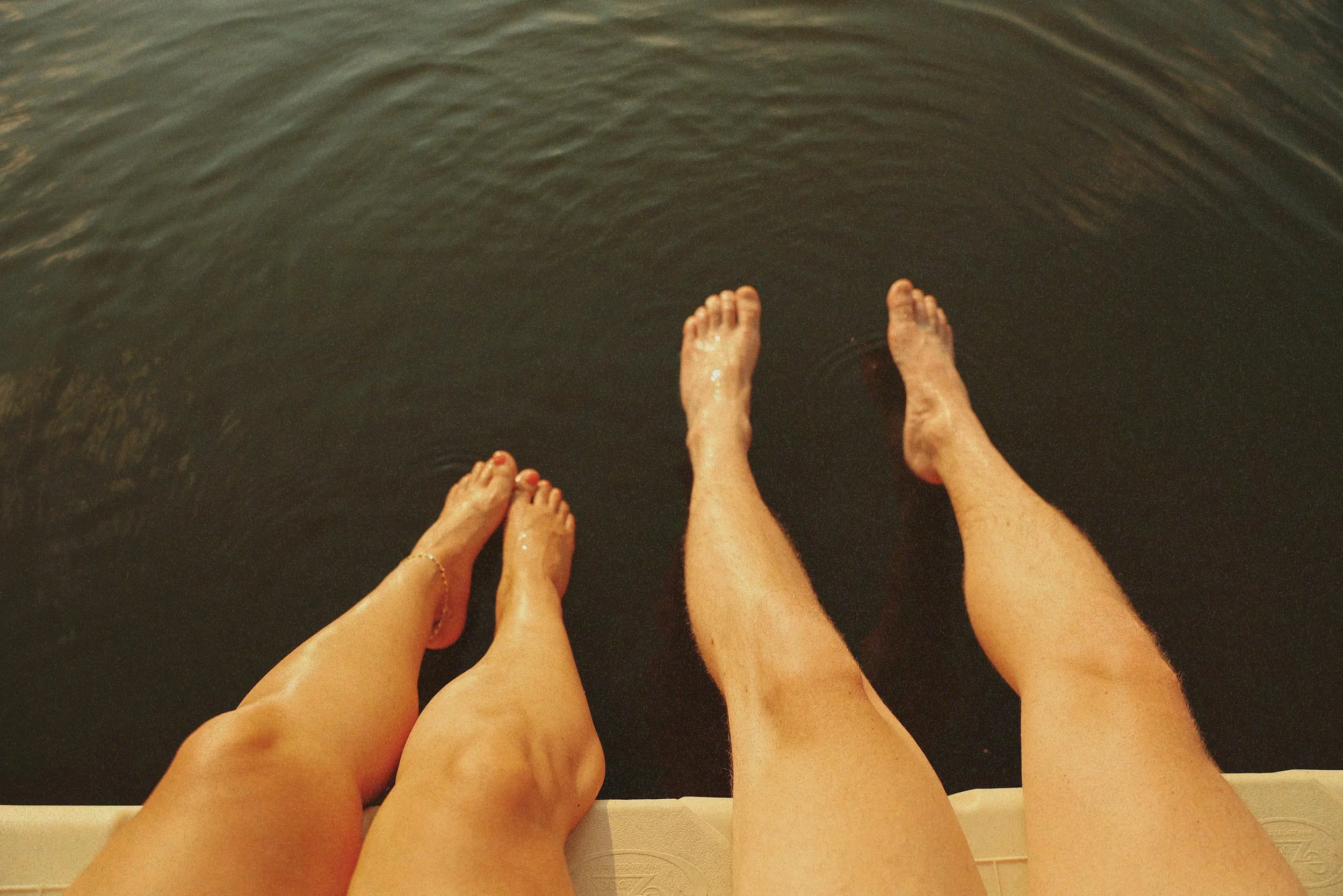 Two pairs of legs and feet are hanging over the edge of a boat, with water below.