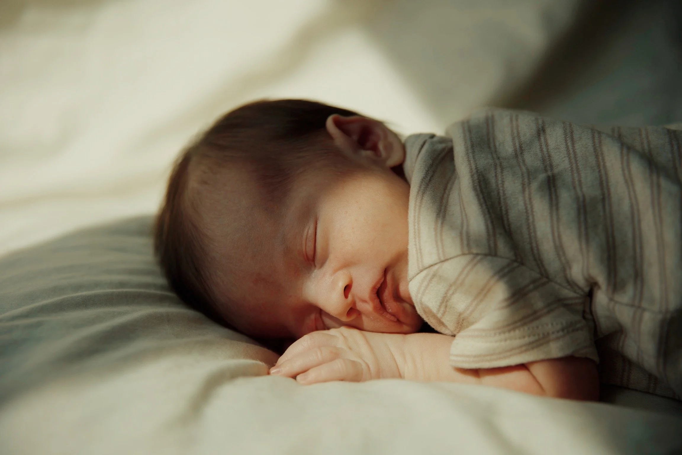 A baby sleeping peacefully on a bed with a soft, cream-colored sheet, dressed in a beige and brown striped onesie.