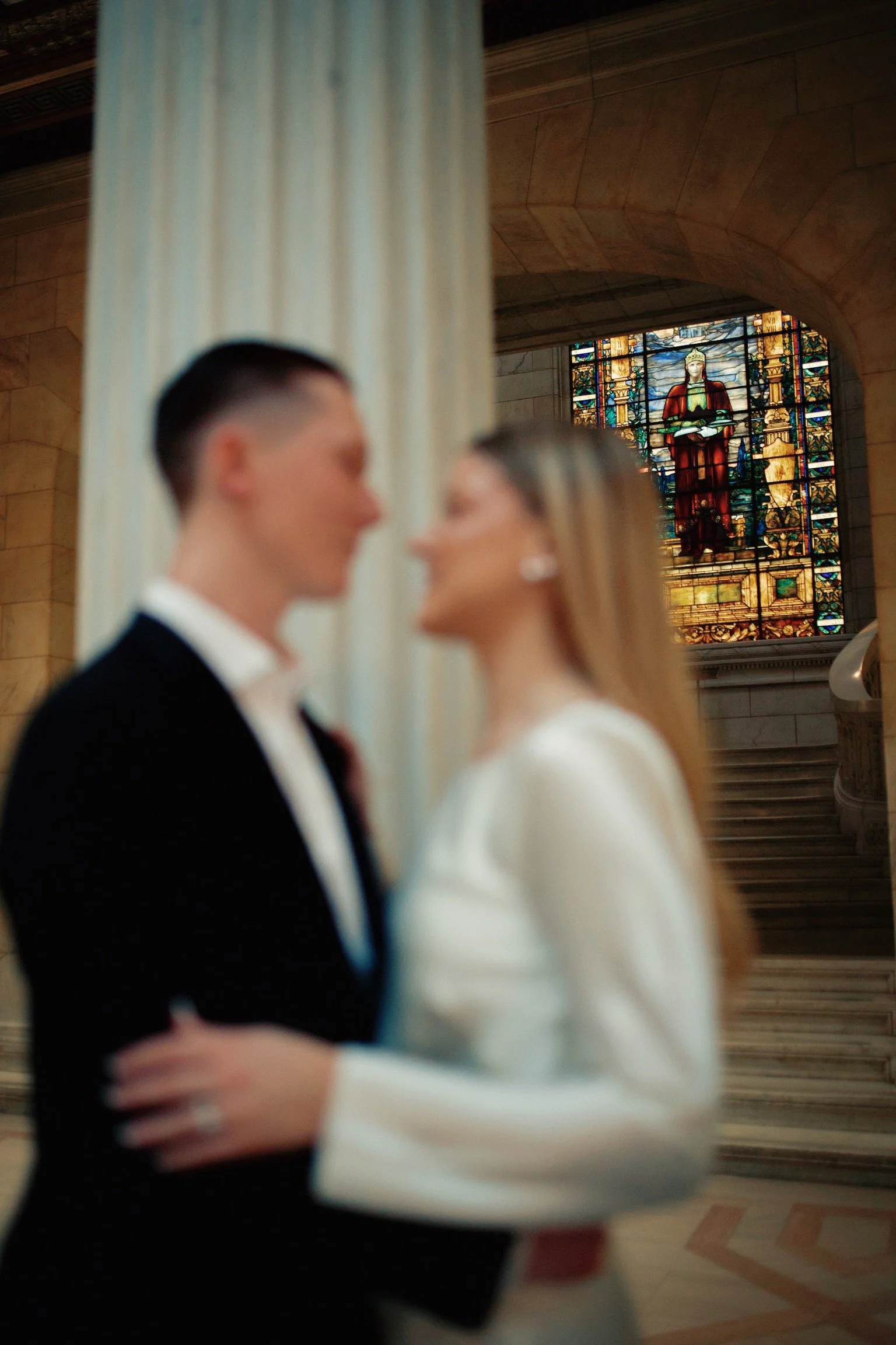Blurred image of a couple in wedding attire embracing in a church interior with a stained glass window in the background.