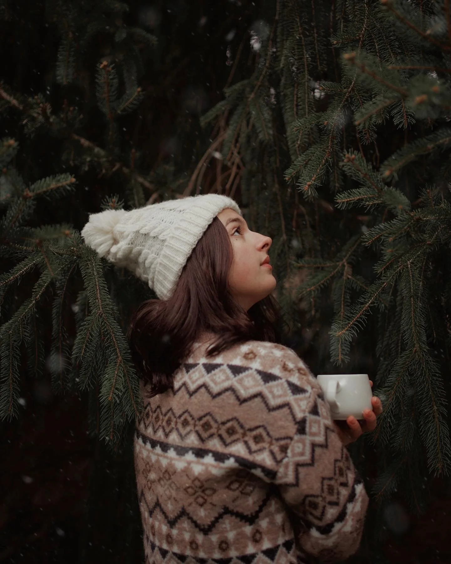 A woman with shoulder-length brown hair wearing a white knitted beanie and a patterned sweater, holding a white mug, standing among evergreen trees during winter.
