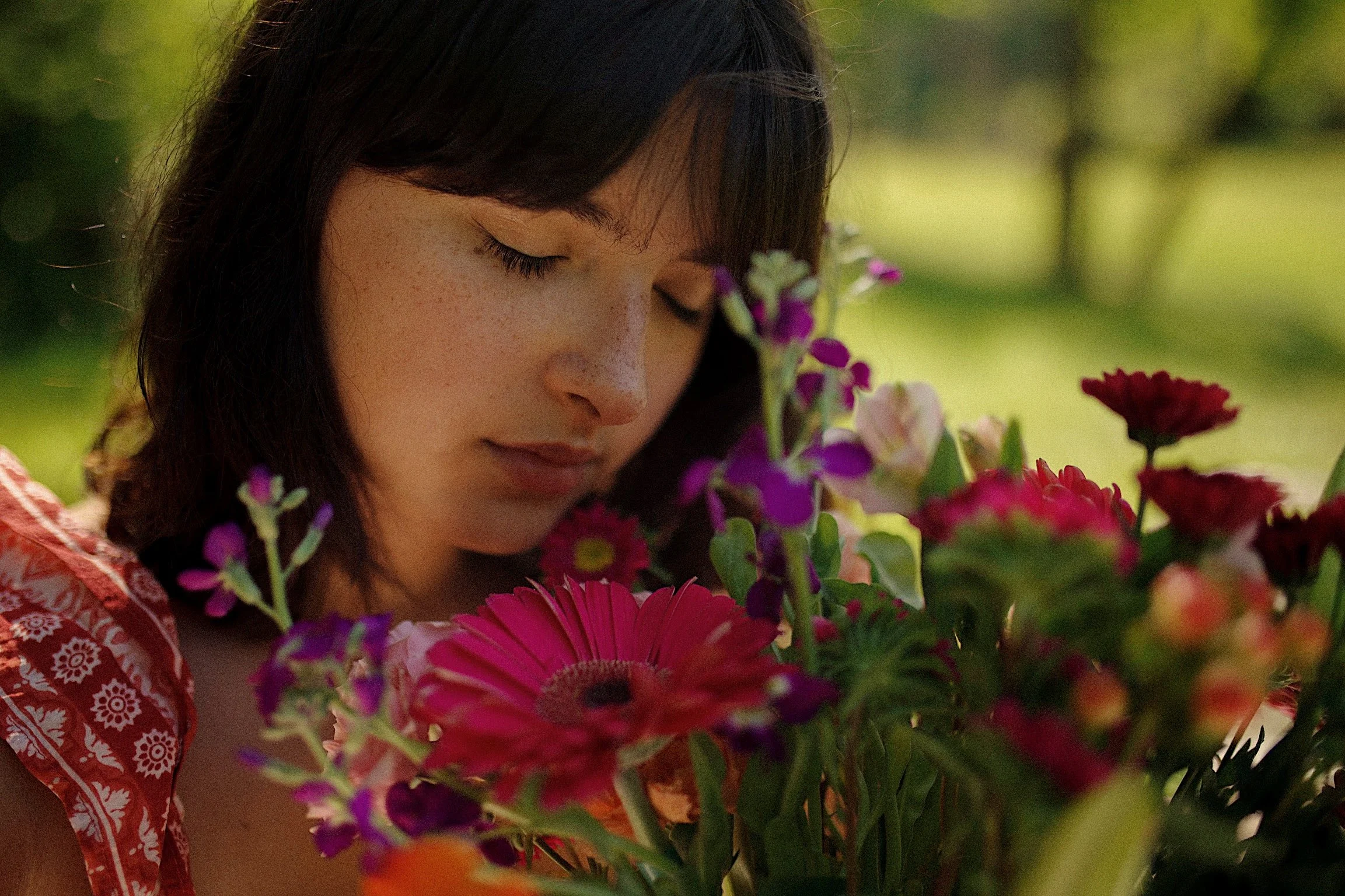 A woman with dark hair and freckles, wearing a patterned orange and white top, gently smelling a bouquet of colorful flowers outdoors during the daytime.