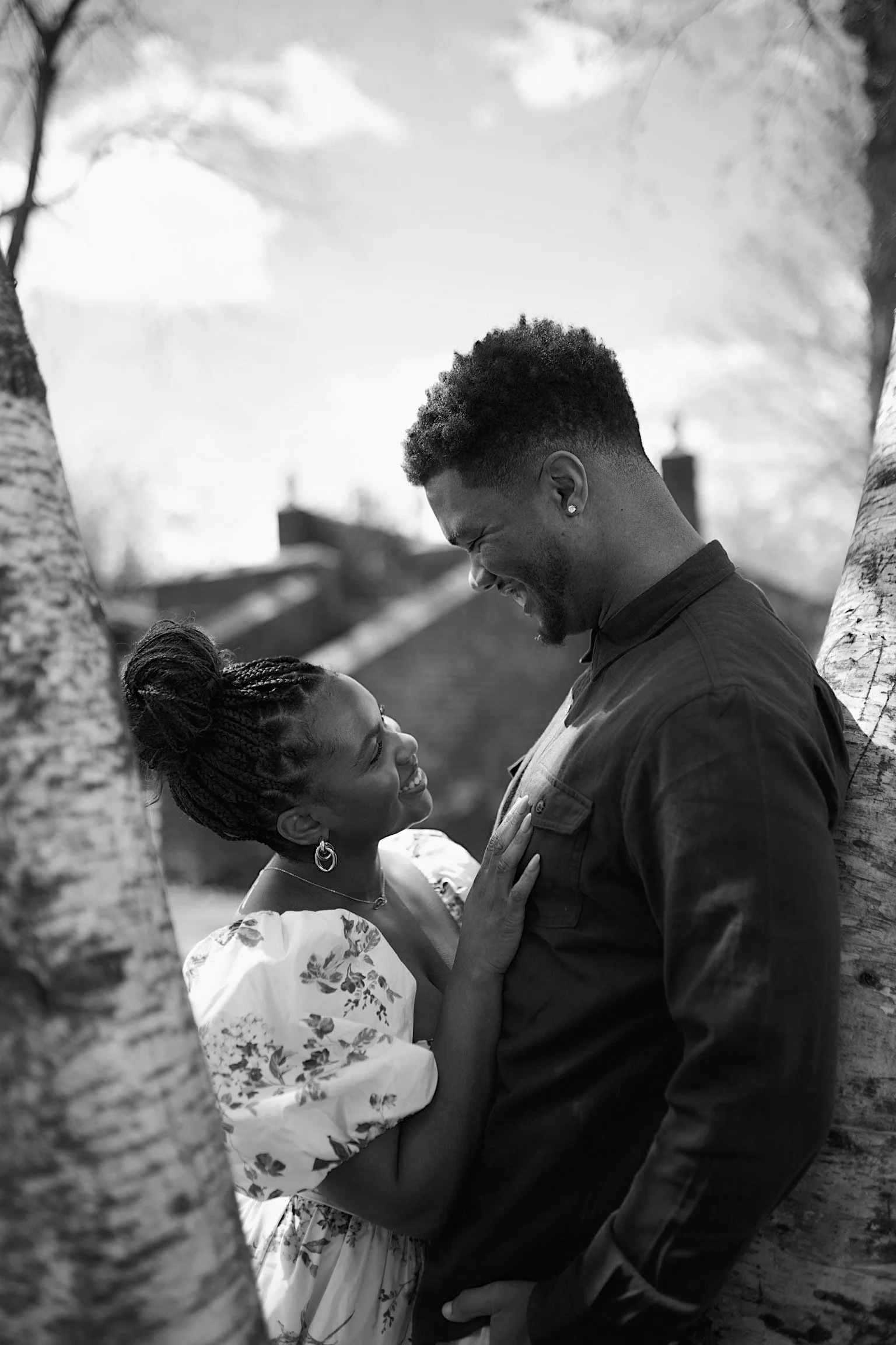 A black and white photo of a couple smiling and looking into each other's eyes, standing between two trees outdoors.