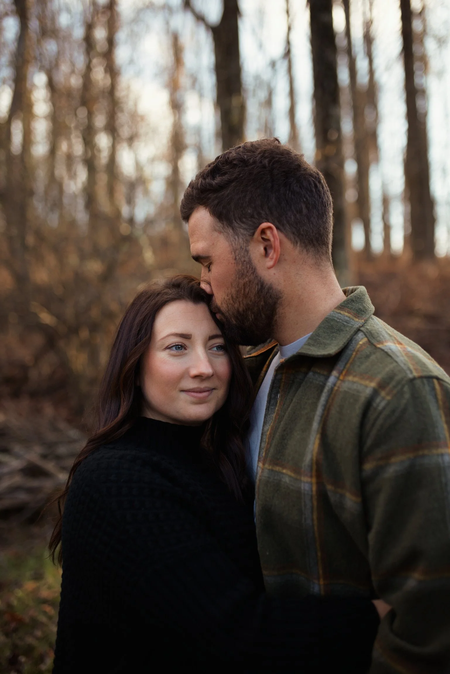 A man with a beard kisses a woman on the forehead in an outdoor wooded area during daytime.