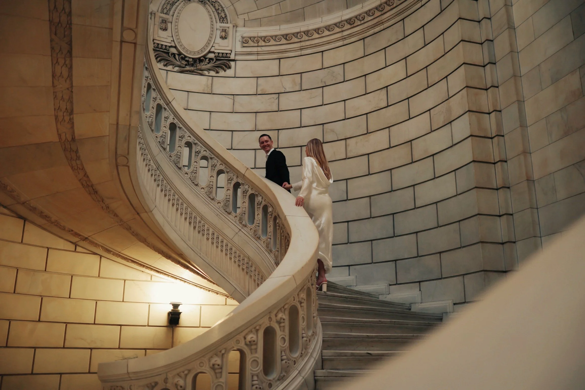 A couple in formal attire ascending a grand, curved stone staircase inside a building with ornate architectural details.