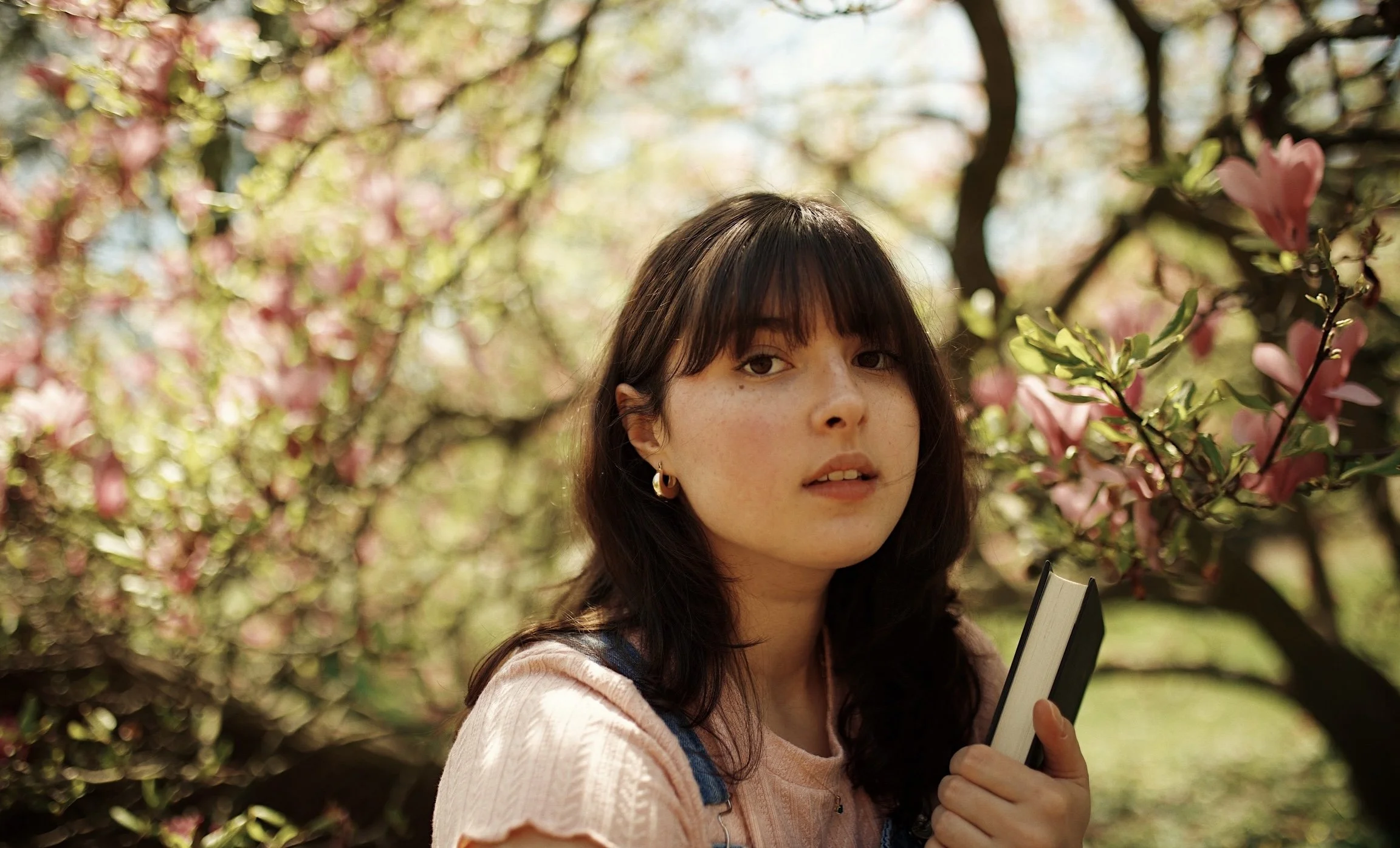 A young woman with dark hair and bangs holding a small notebook in a garden with blooming pink and white flowers on trees.