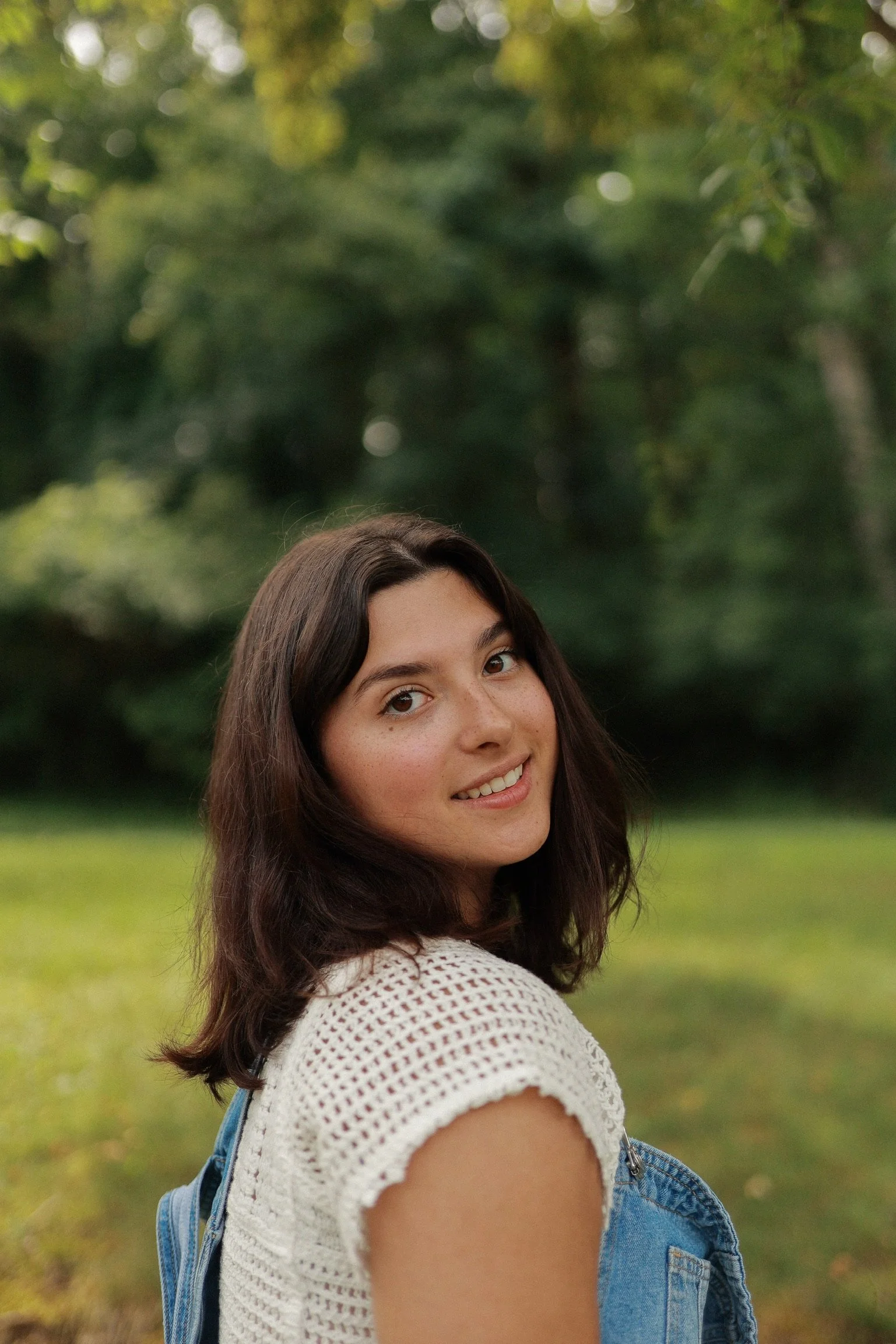 A young woman with dark brown hair and freckles smiling outdoors with a green, leafy background.