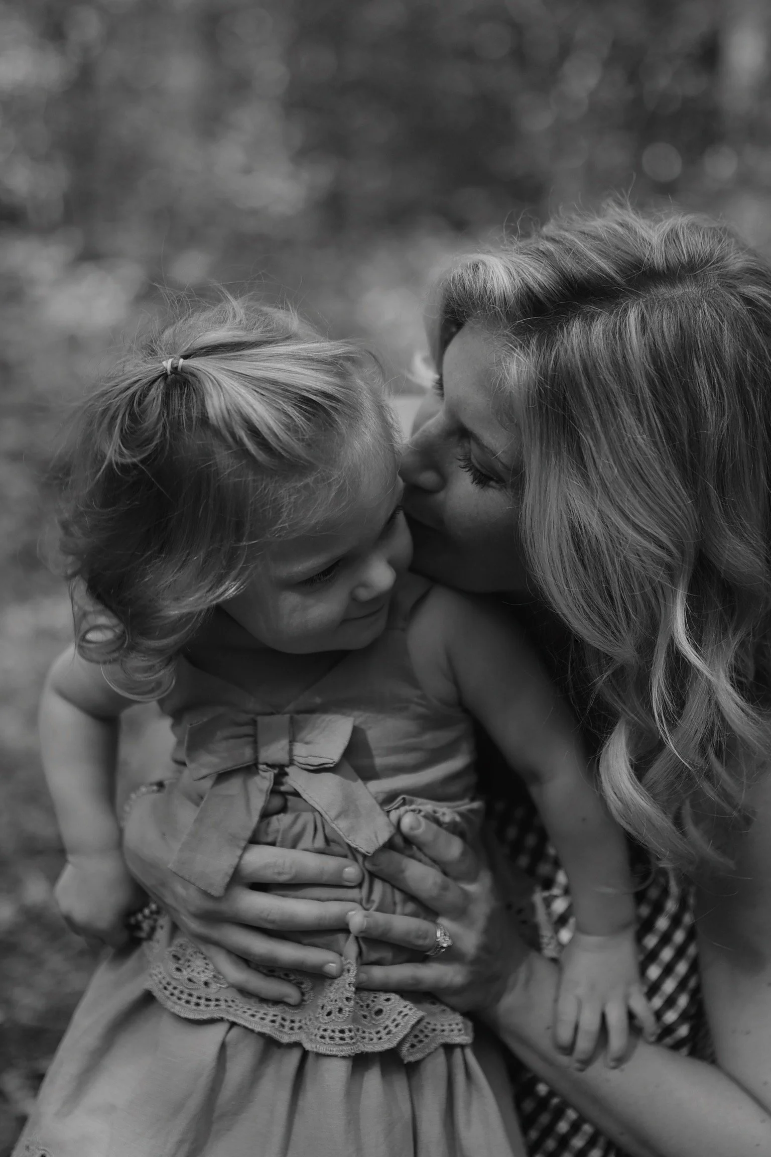 A woman is giving a gentle kiss on the forehead of a young girl, with their faces close together. The girl has curly hair and is wearing a dress with a bow. The woman has long, wavy hair and is holding the girl with both hands.