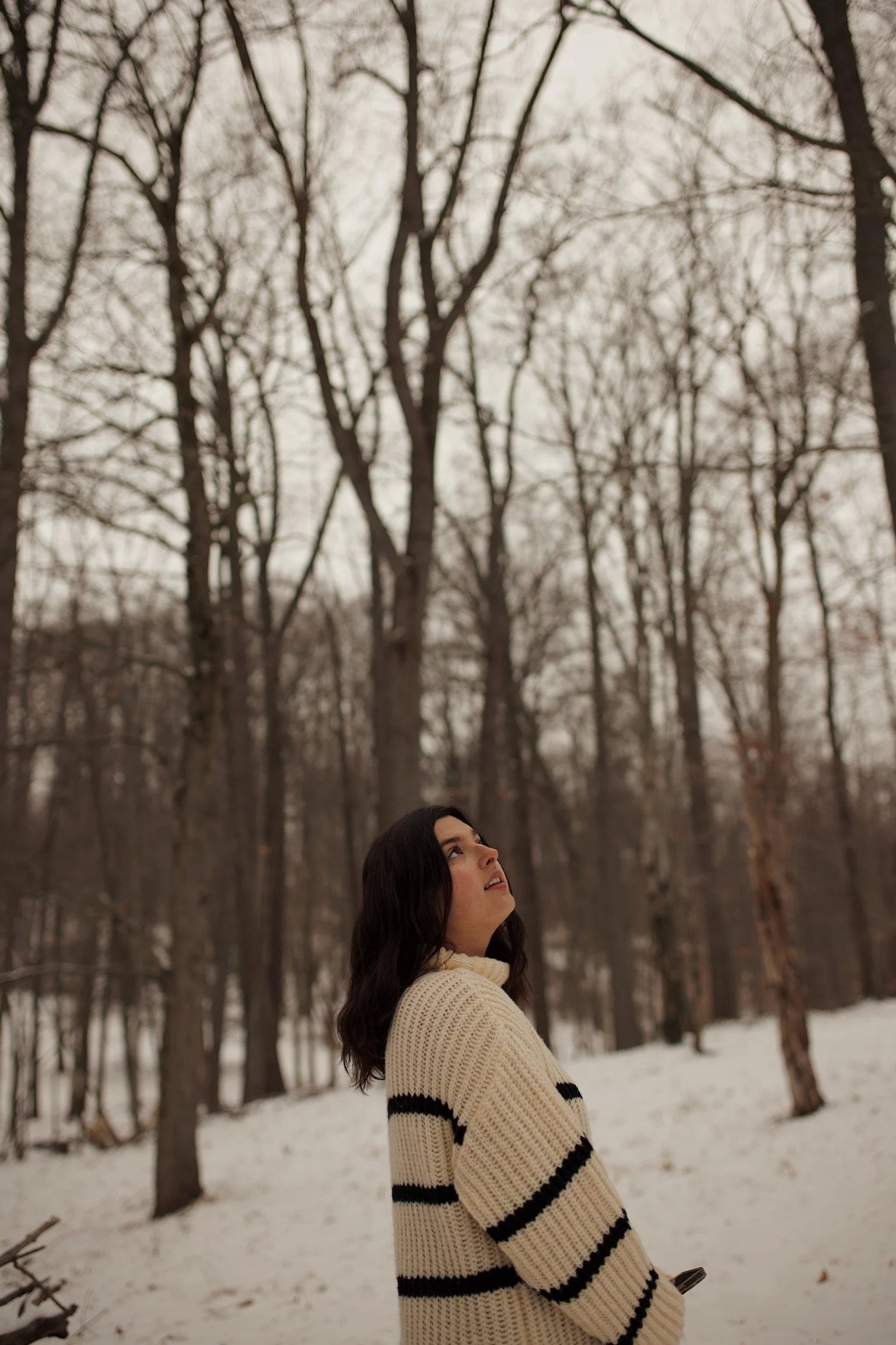 A woman with dark hair wearing a cream-colored knit sweater with black stripes standing in a snowy forest, looking up at the leafless trees.