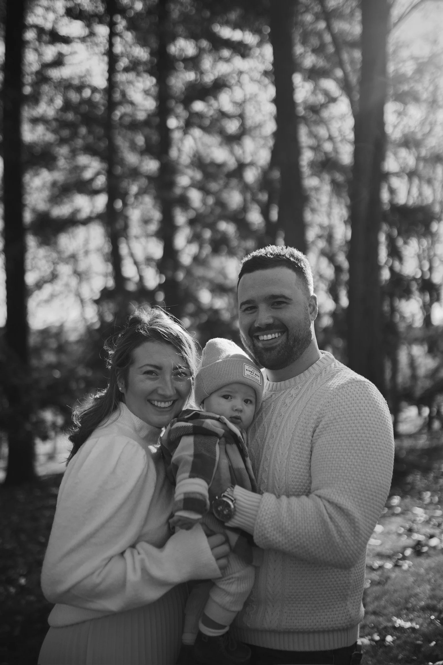 Black and white photo of a smiling family of three outdoors in a forest with trees, showing a woman, man, and a baby.