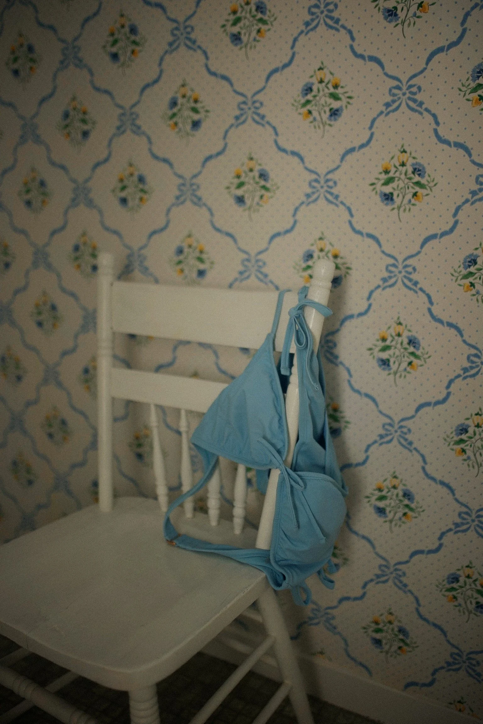 A white wooden chair with a light blue backpack hanging on its backrest, set against a wallpapered wall with a blue, yellow, and green floral pattern.