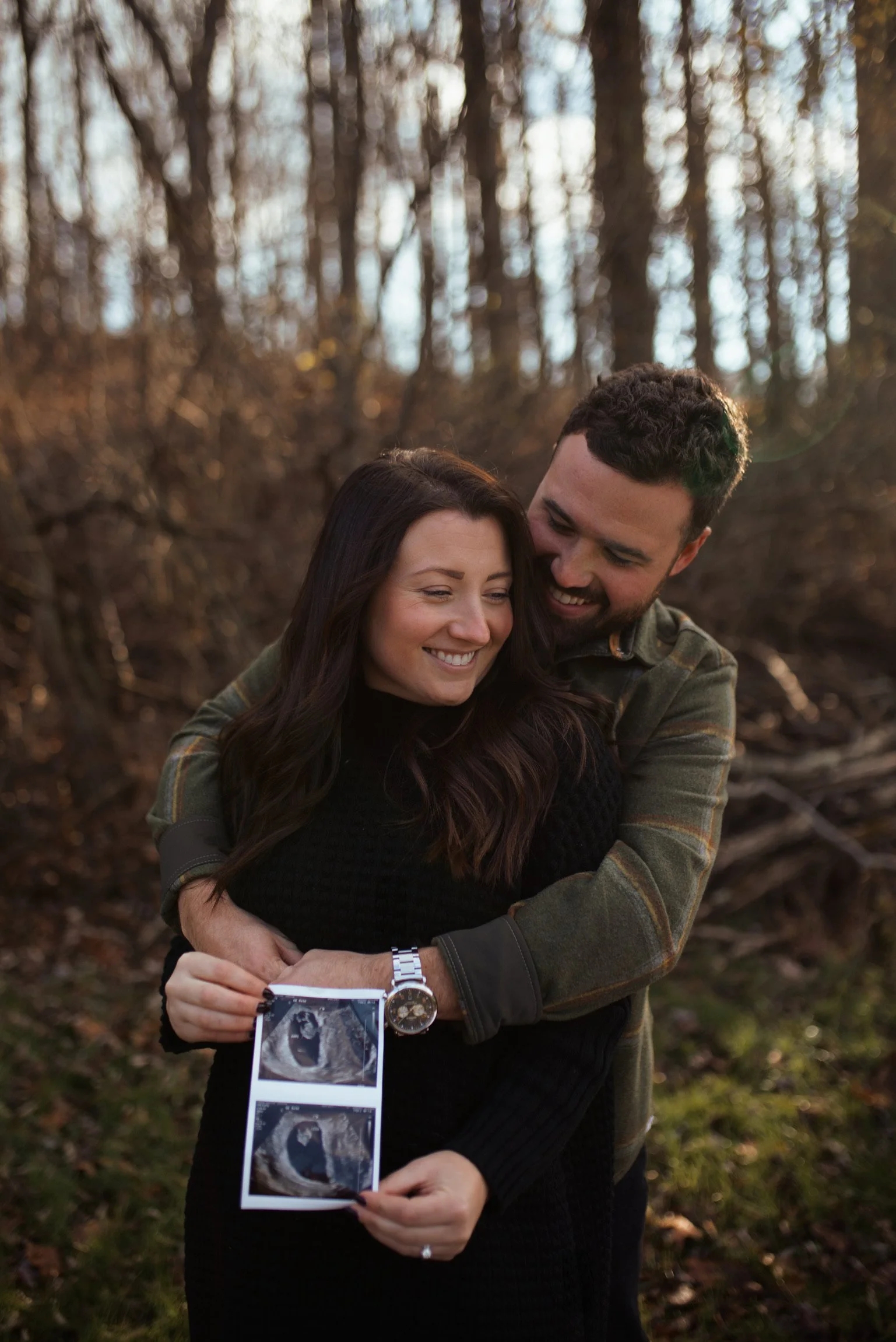 A couple hugging outdoors in a wooded area, holding ultrasound pictures, smiling.