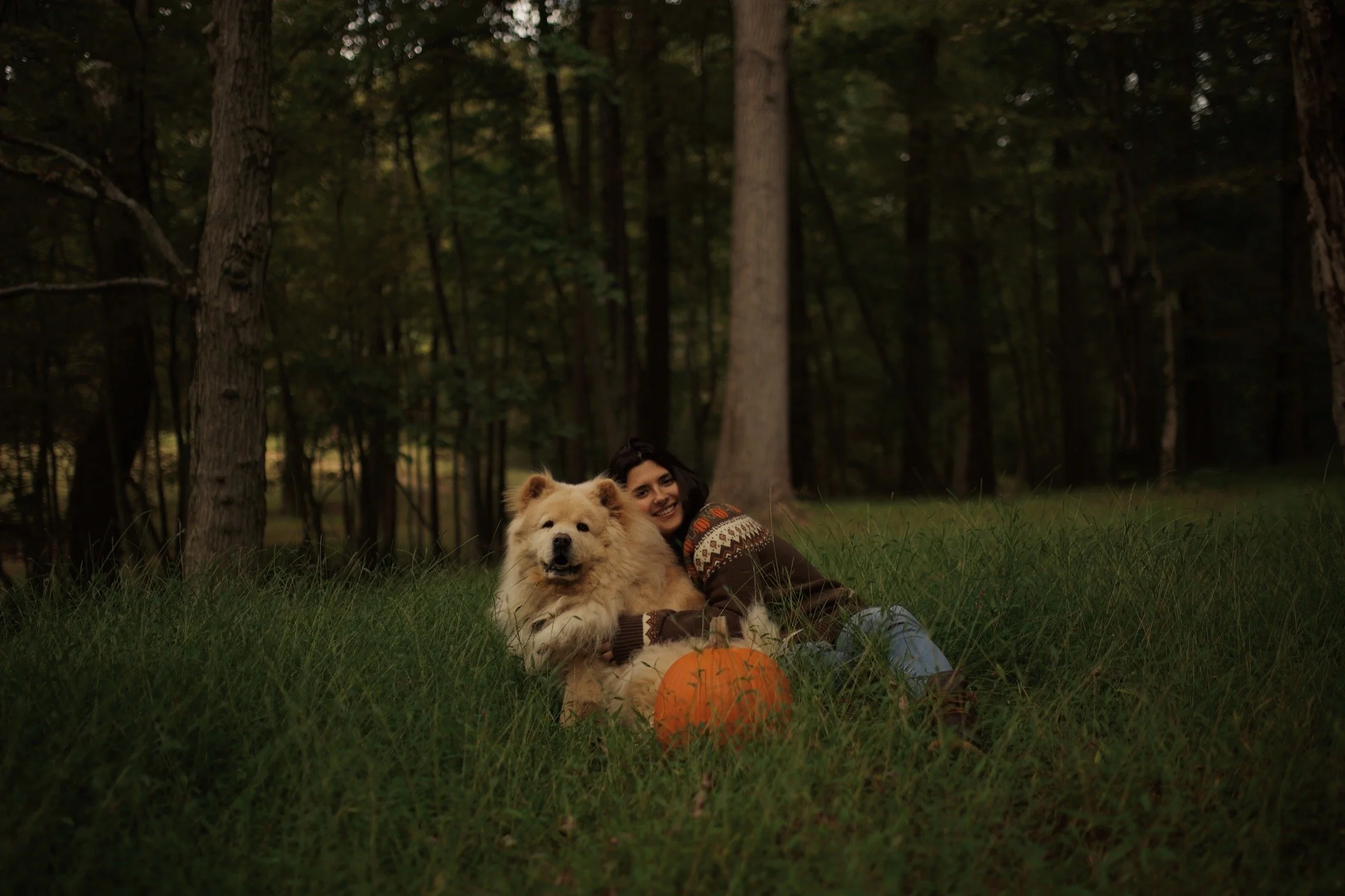 A woman and a large fluffy dog sitting on grass in a wooded area with trees, smiling, with a pumpkin placed in front of the dog.
